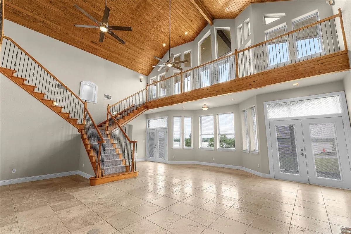 Spacious living area featuring a vaulted wood-paneled ceiling with ceiling fans, a grand bifurcated staircase with wood treads and wrought iron balusters, tile flooring, and multiple windows with outdoor scenery
