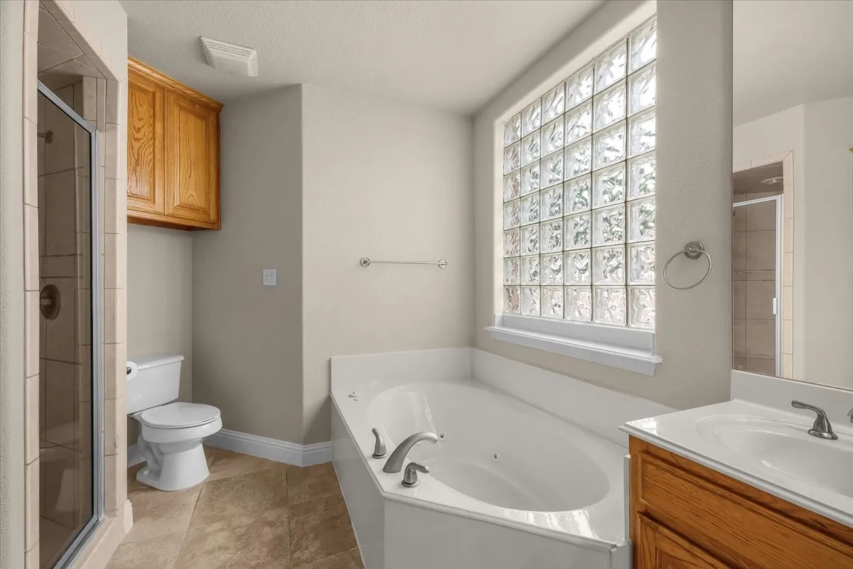 Bathroom featuring a corner soaking tub, a large glass block window, a glass-enclosed shower, a wood-finish vanity with an integrated sink, and floor tiling