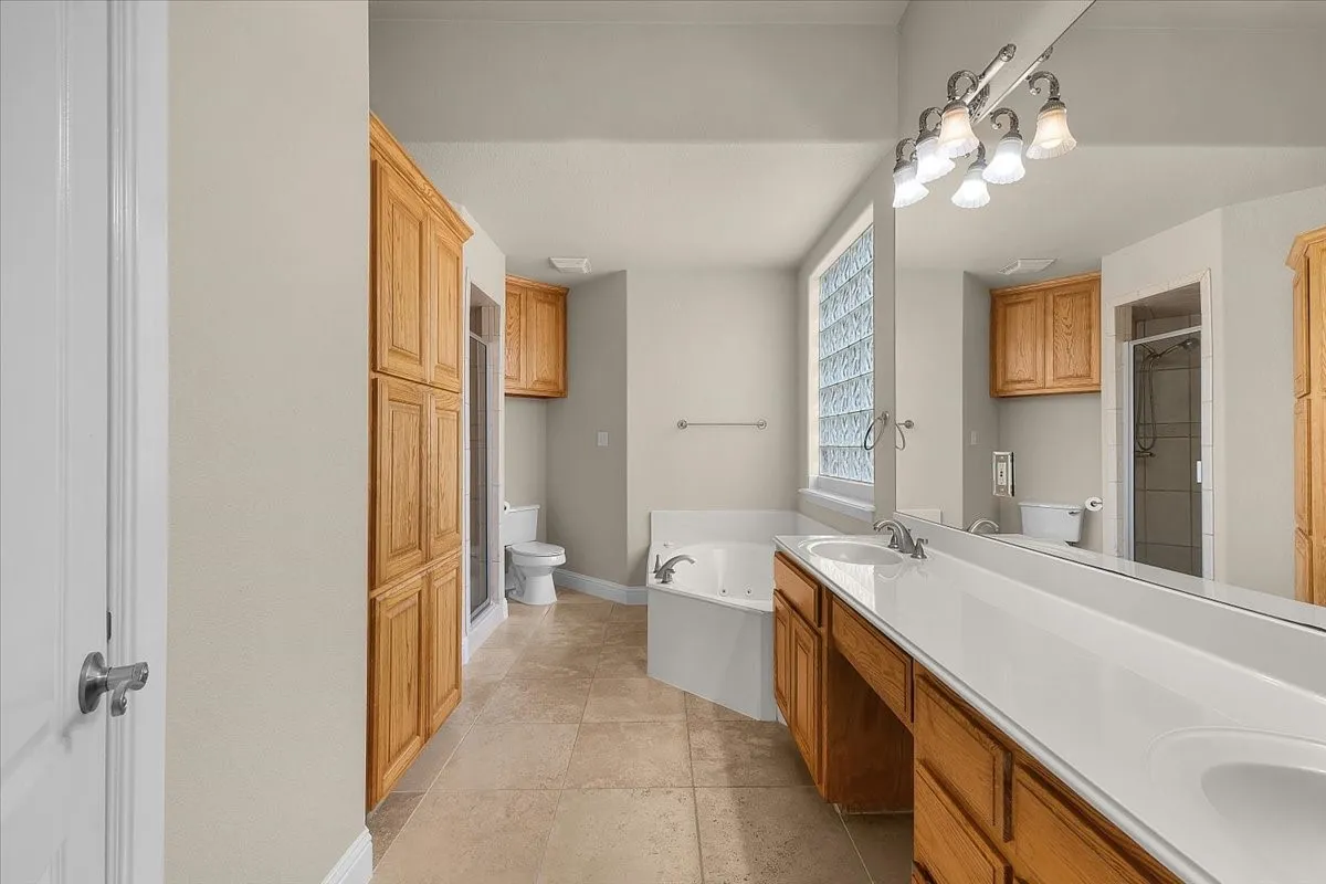 Dual vanity with integrated sinks and wood cabinetry, alongside a jetted tub, glass block window, and tile flooring
