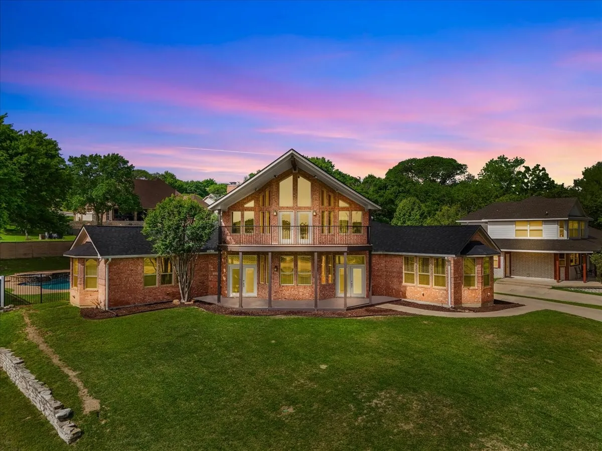 Brick facade home with a prominent two-story window wall, second-story balcony, covered patio, expansive green lawn, and a fenced swimming pool