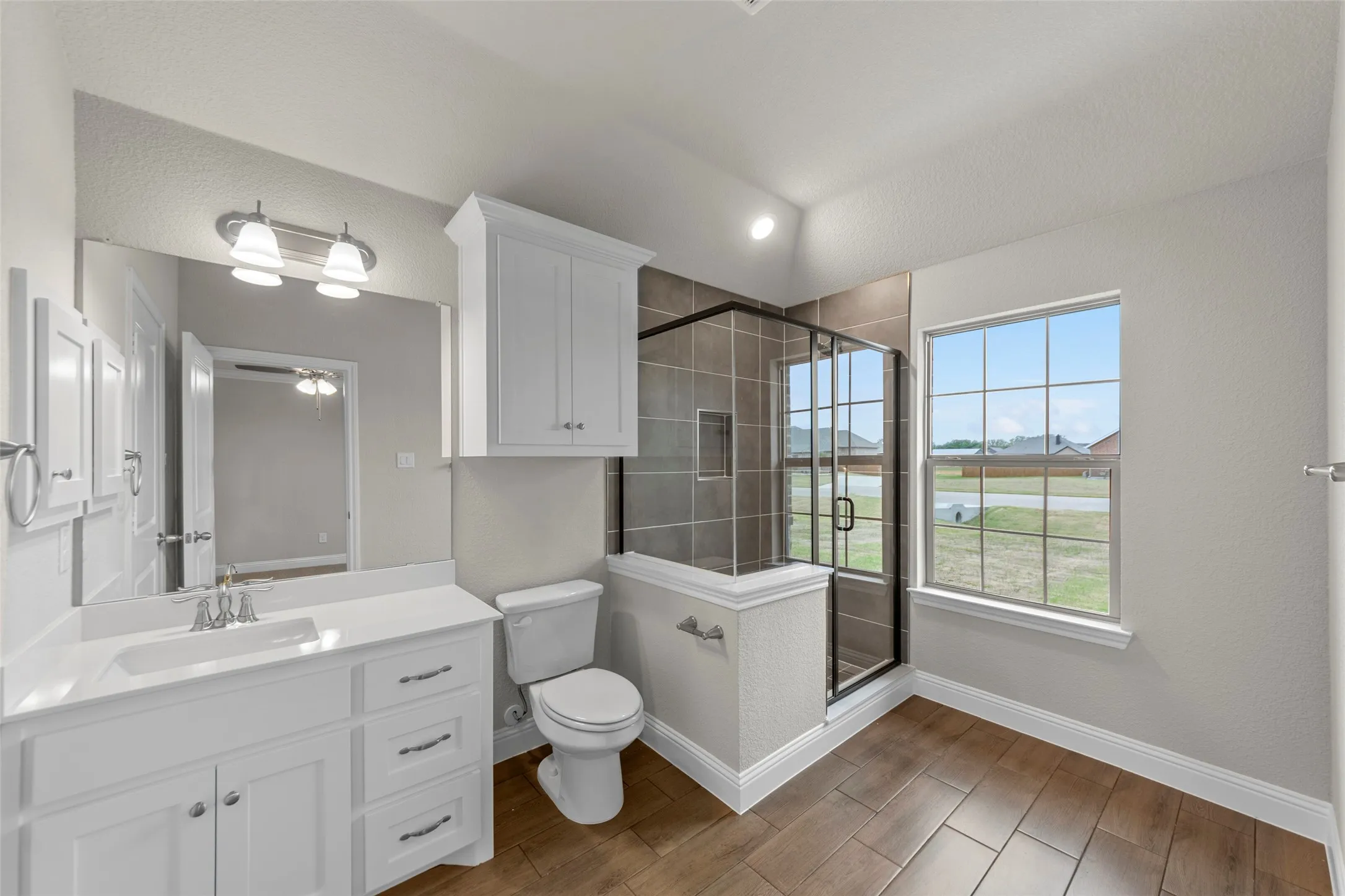 Bathroom featuring a single vanity with white cabinetry, a walk-in shower with dark-toned tile surround, wood-finish flooring, a toilet, and an overhead storage cabinet