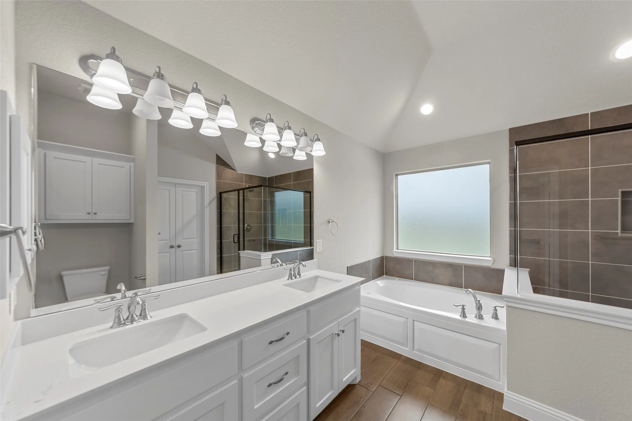 Spacious bathroom featuring a dual vanity with white cabinetry and chrome fixtures, a large soaking tub, a tiled walk-in shower with a glass enclosure, and wood-finish flooring