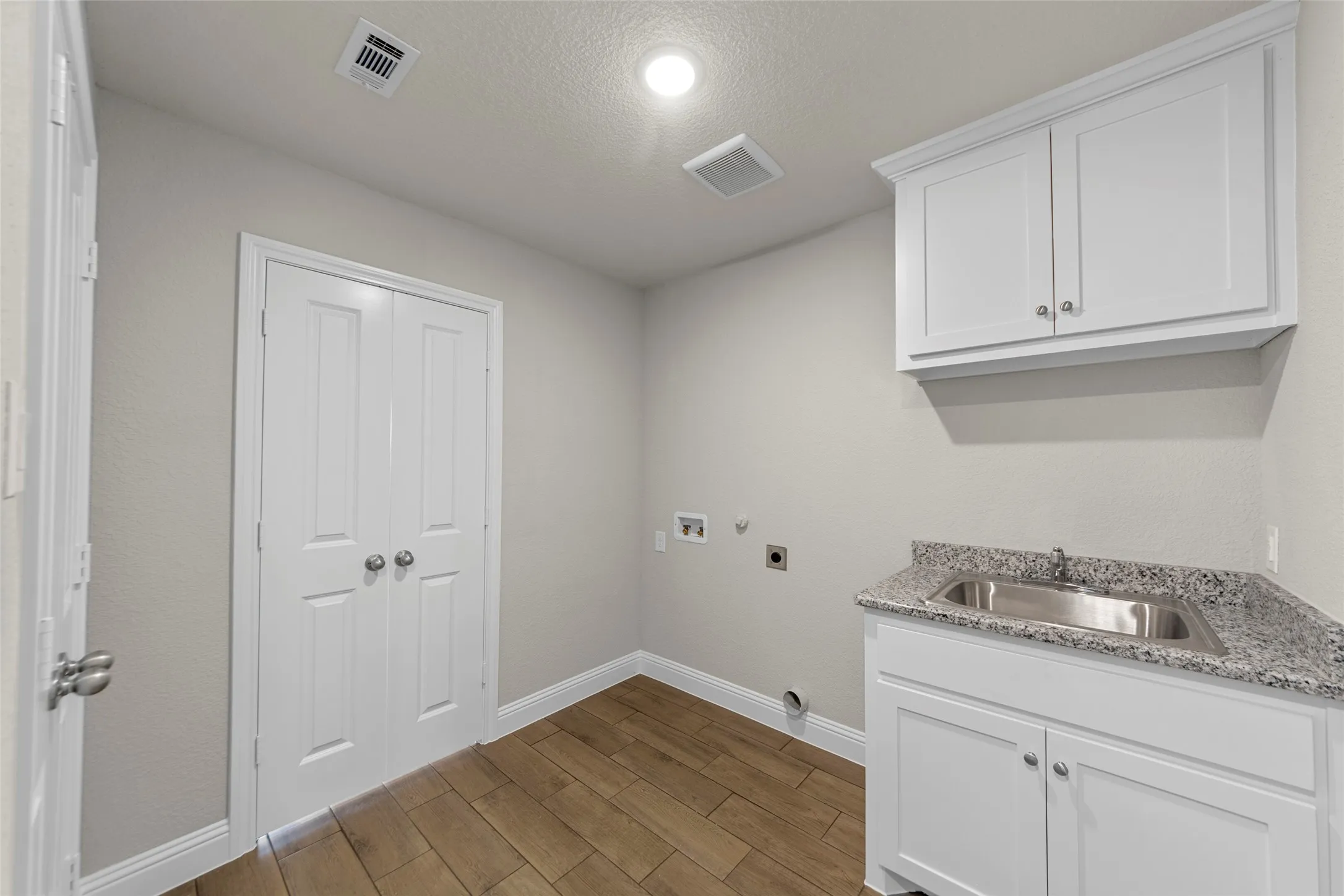 Dedicated utility space featuring wood-finish flooring, white cabinetry, a stainless steel sink with a granite countertop, and bi-fold doors