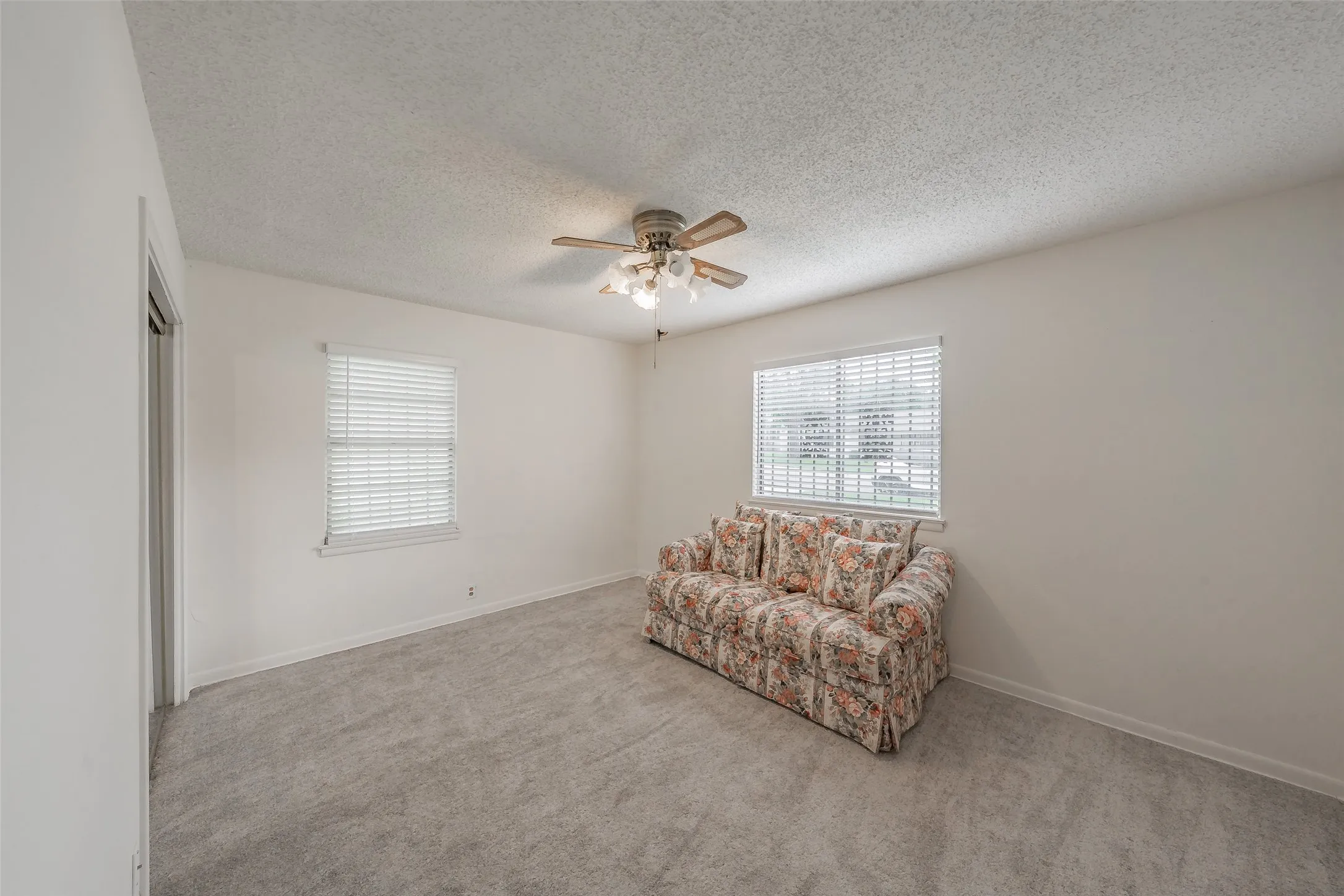 Spacious room featuring neutral carpeting, two windows with horizontal blinds, and a ceiling fan with light fixture