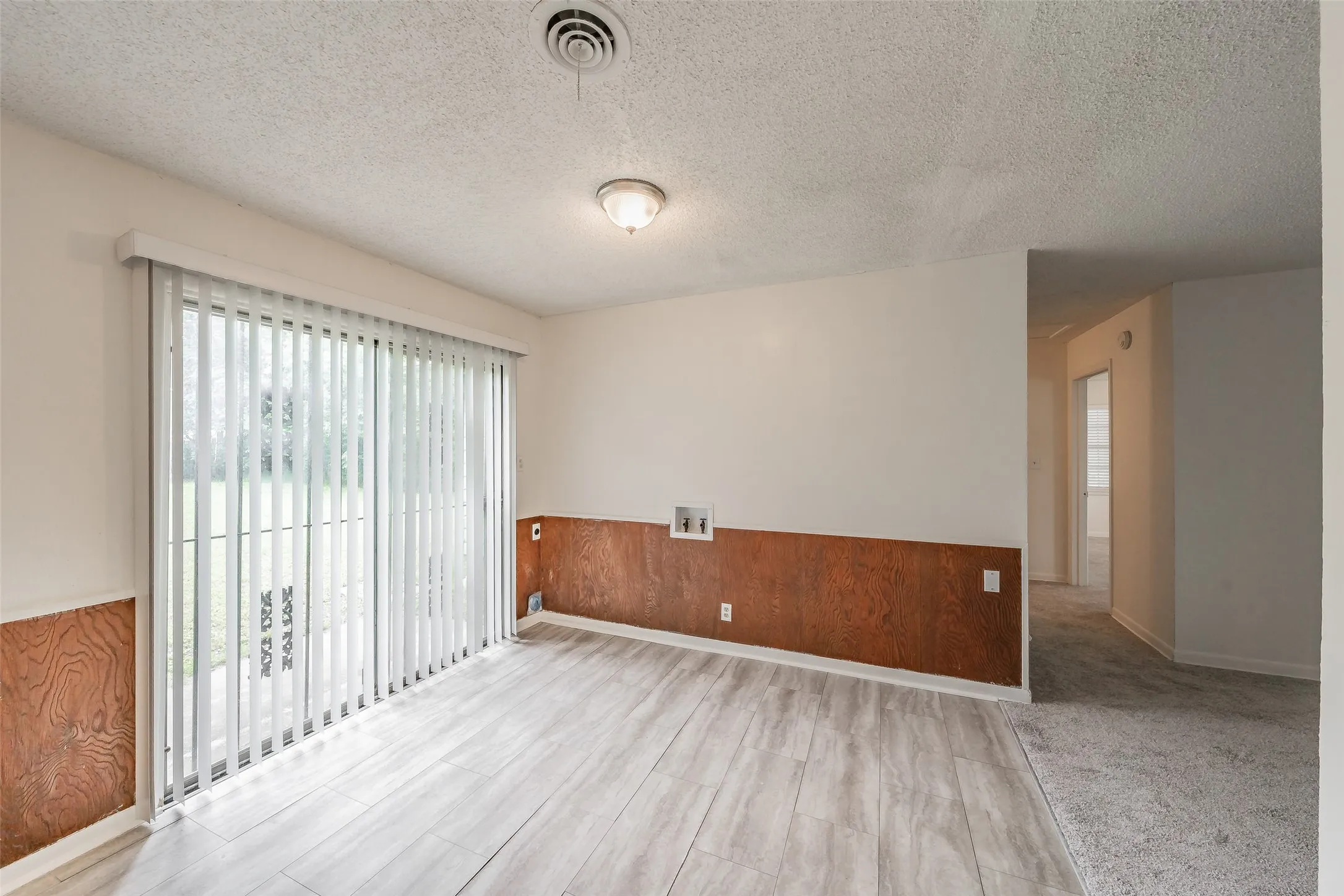 Spacious room featuring light wood-finish flooring, white walls, and a large sliding glass door with vertical blinds