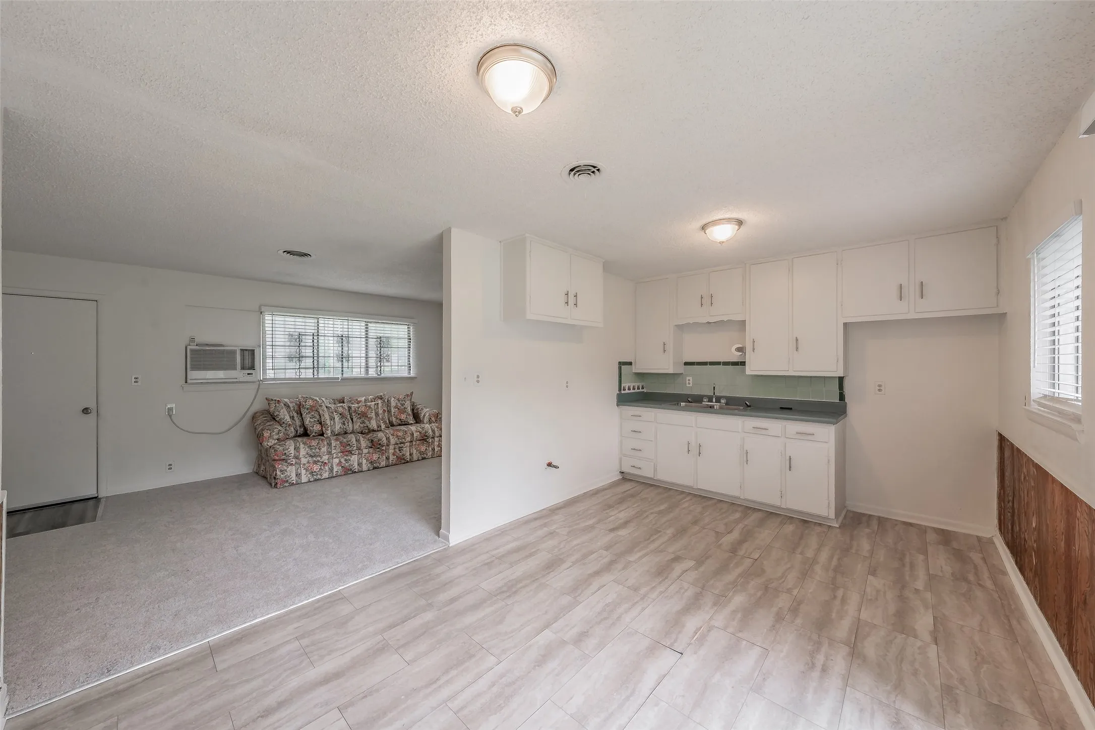 Open-concept living area featuring a kitchen with white cabinetry and a window, and a carpeted room with a window and a wall-mounted air conditioning unit