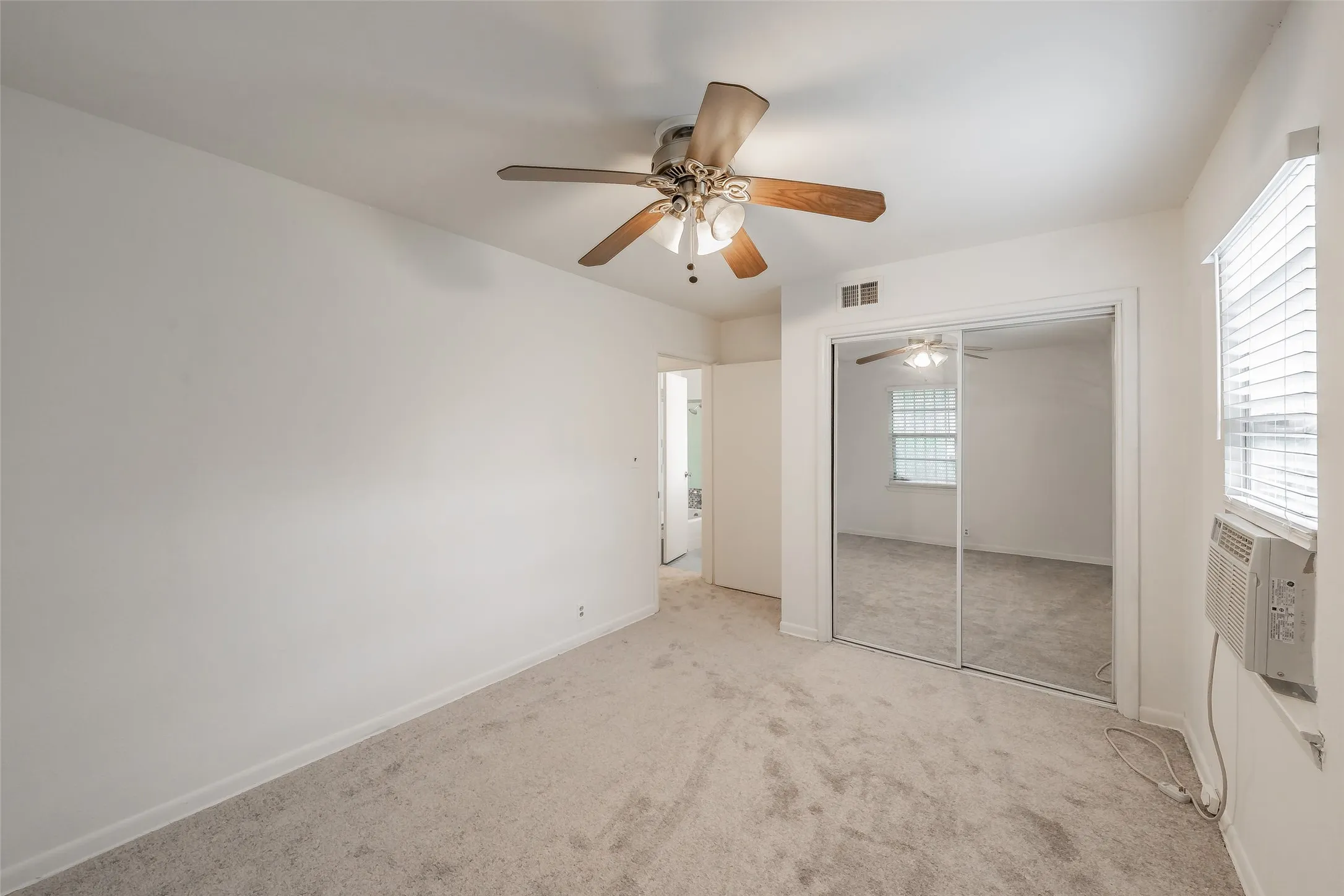 Carpeted room featuring a ceiling fan with light fixture, mirrored sliding closet doors, window blinds, and a wall-mounted air conditioning unit