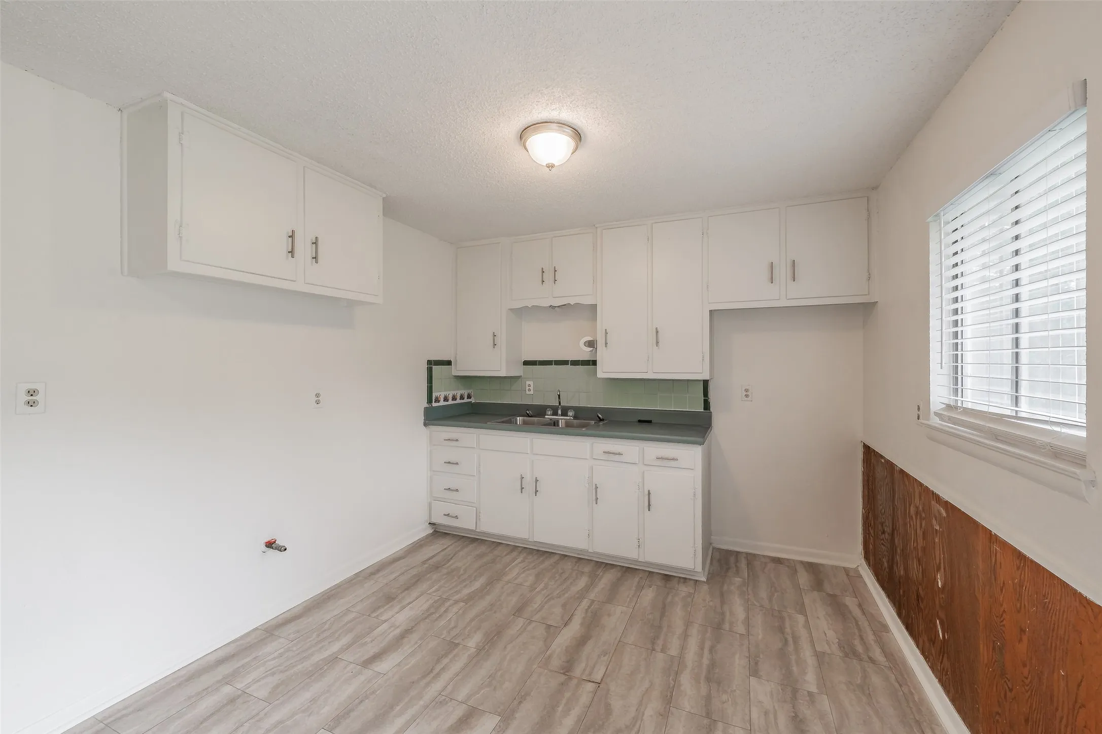 Kitchen area featuring light wood-finish flooring, white cabinetry with silver-tone hardware, a sink with a dark countertop, a light green tiled backsplash, and a window with blinds