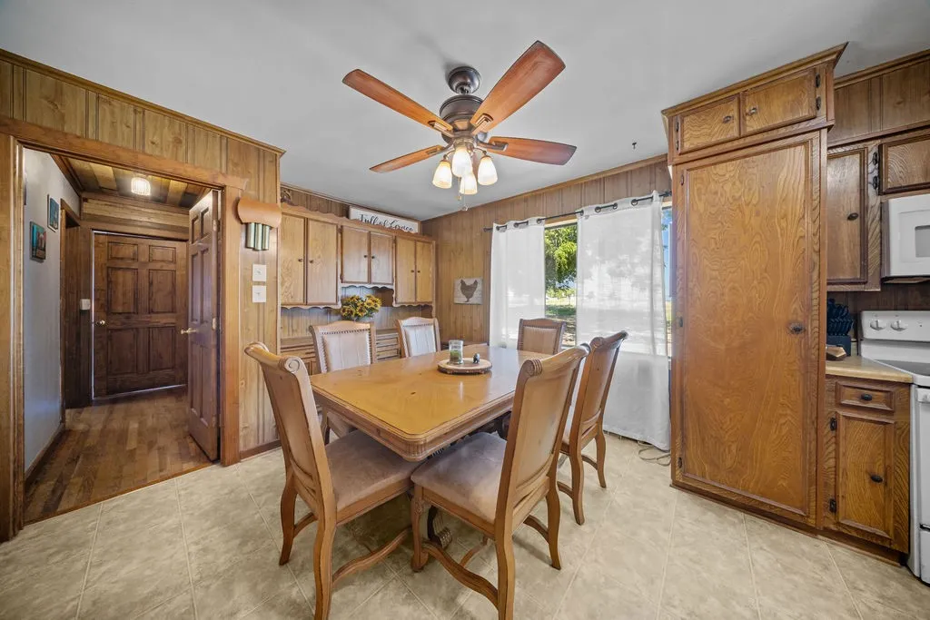 Dining area featuring wood paneling, a ceiling fan with light fixture, and a window with white curtains