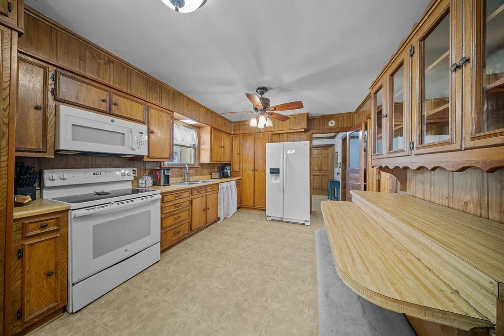 The kitchen features wood paneling, a ceiling fan with lighting, white appliances, and ample cabinetry, including some with glass fronts