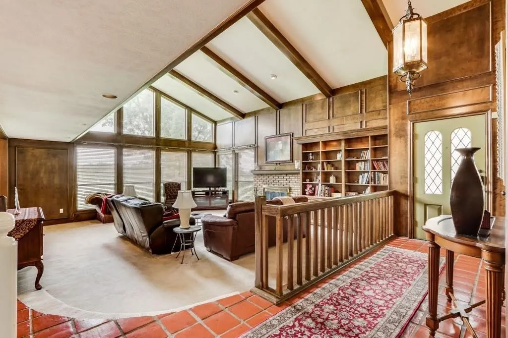 Vaulted ceiling with exposed wood beams, expansive window wall, built-in shelving, wood wall paneling, and terra cotta floor tiles