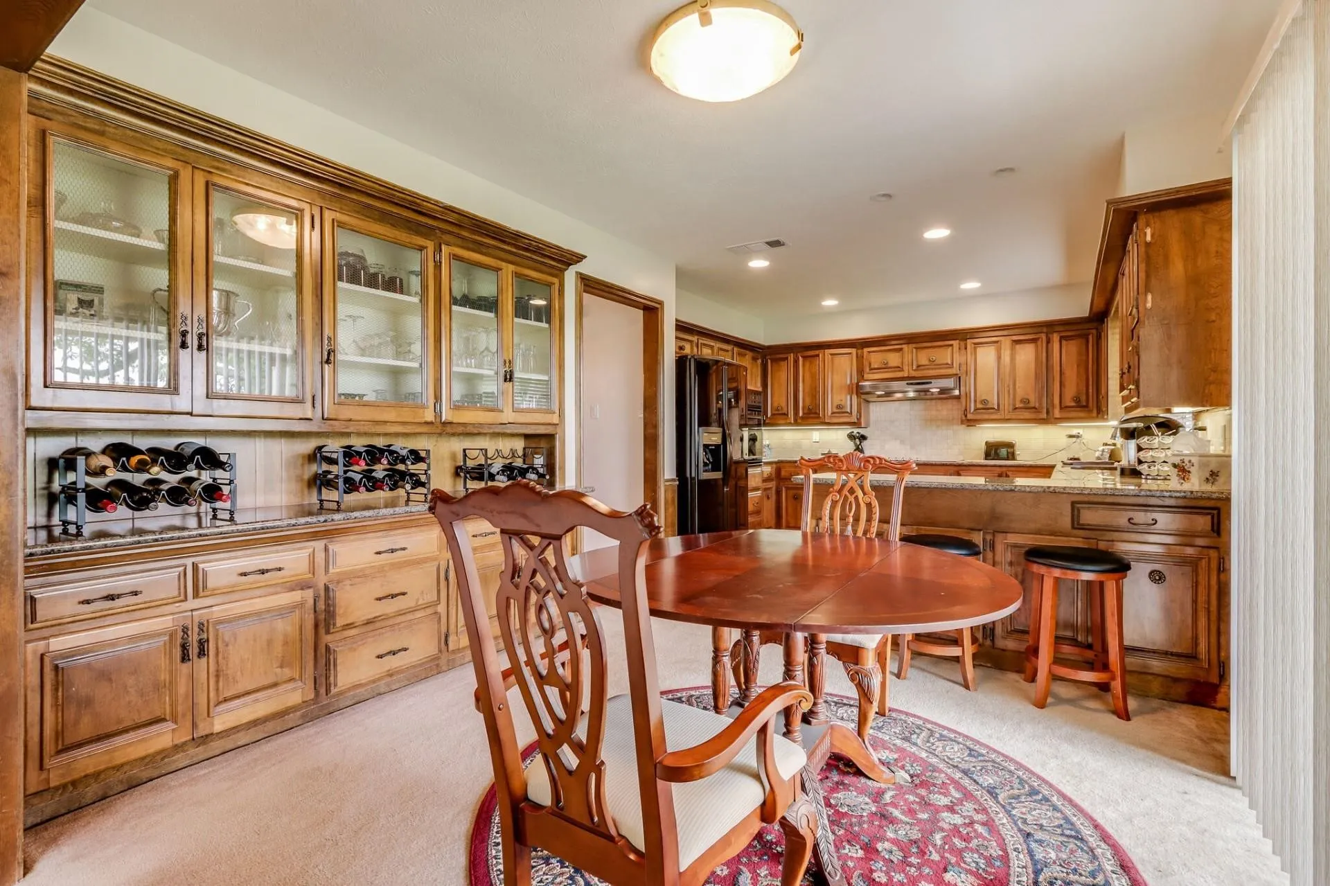 Kitchen dining area with a circular wood-finish table, adjacent to a full kitchen featuring wood-finish cabinetry, a tile backsplash, and recessed lighting