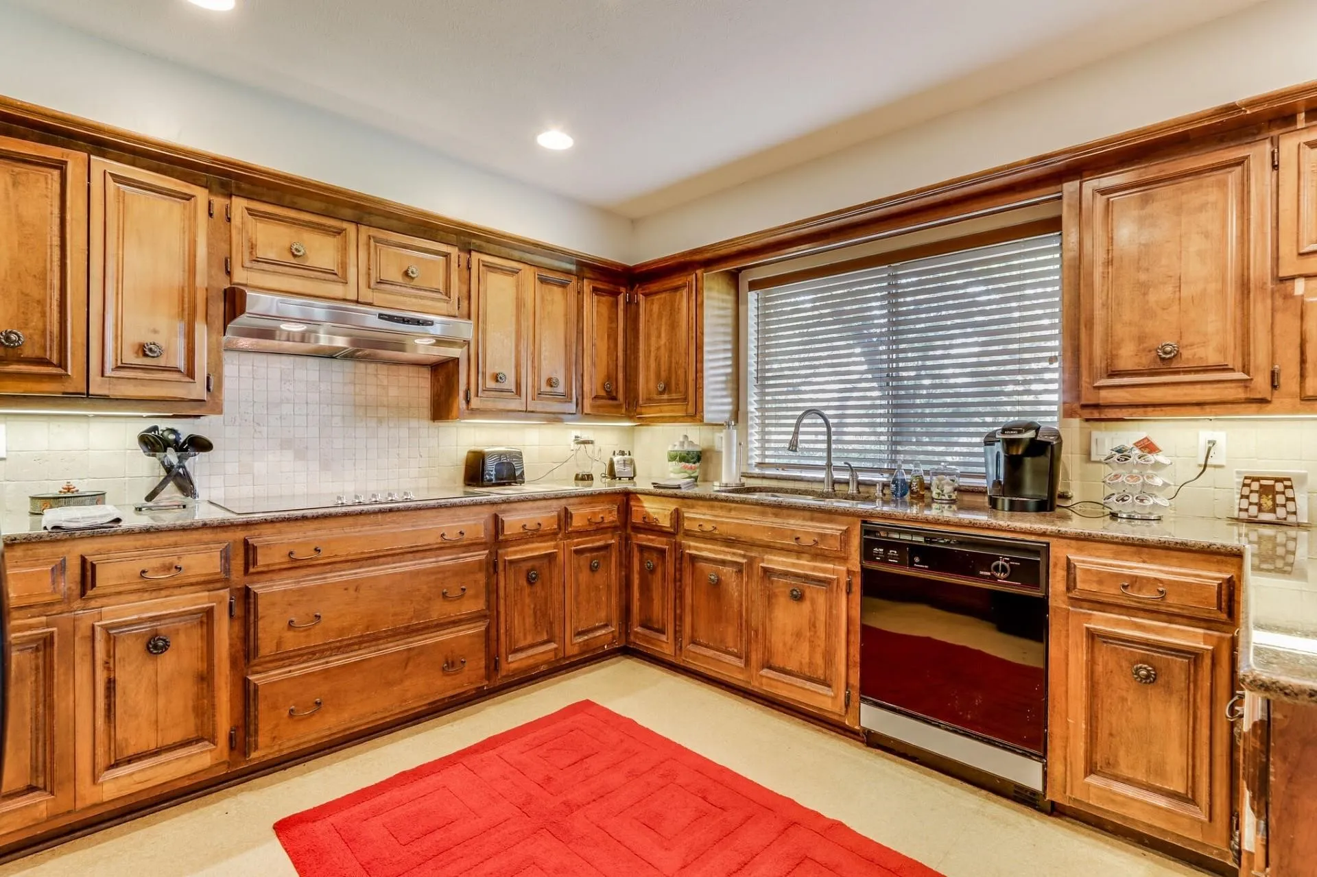 Kitchen featuring wood cabinetry, light-toned countertops, tile backsplash, stainless steel range hood, and a built-in dishwasher
