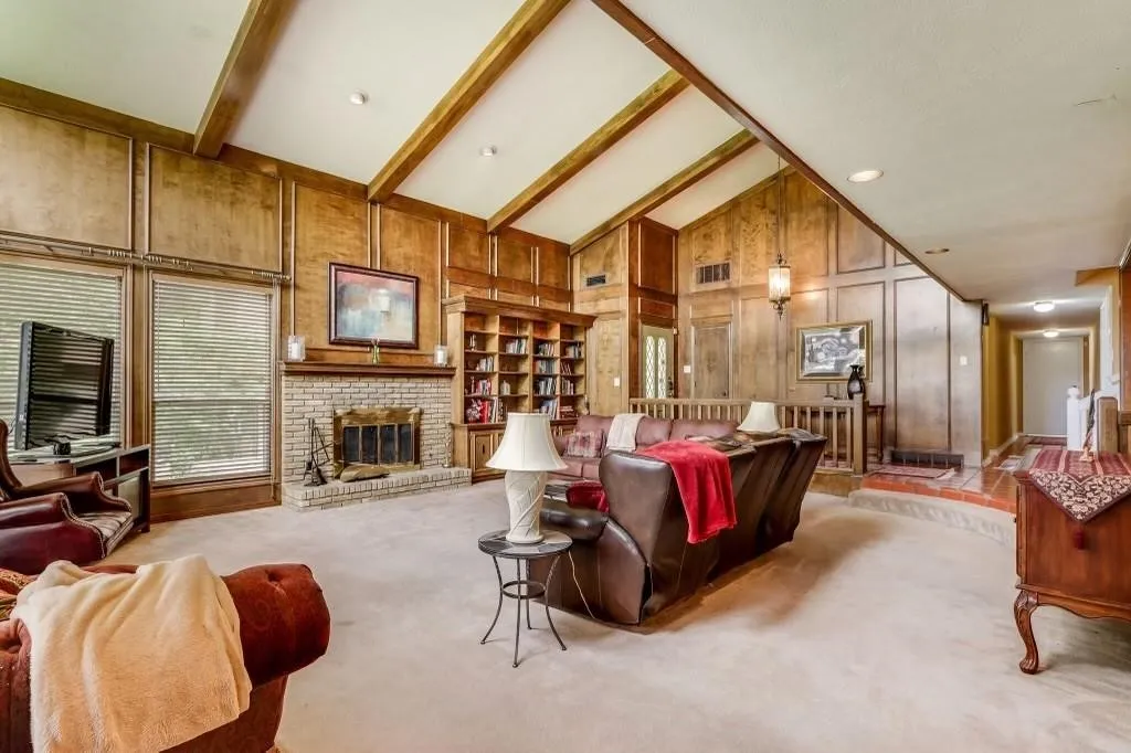 Spacious living area featuring a vaulted ceiling with exposed wood beams, extensive wood paneling, a brick fireplace, and built-in shelving