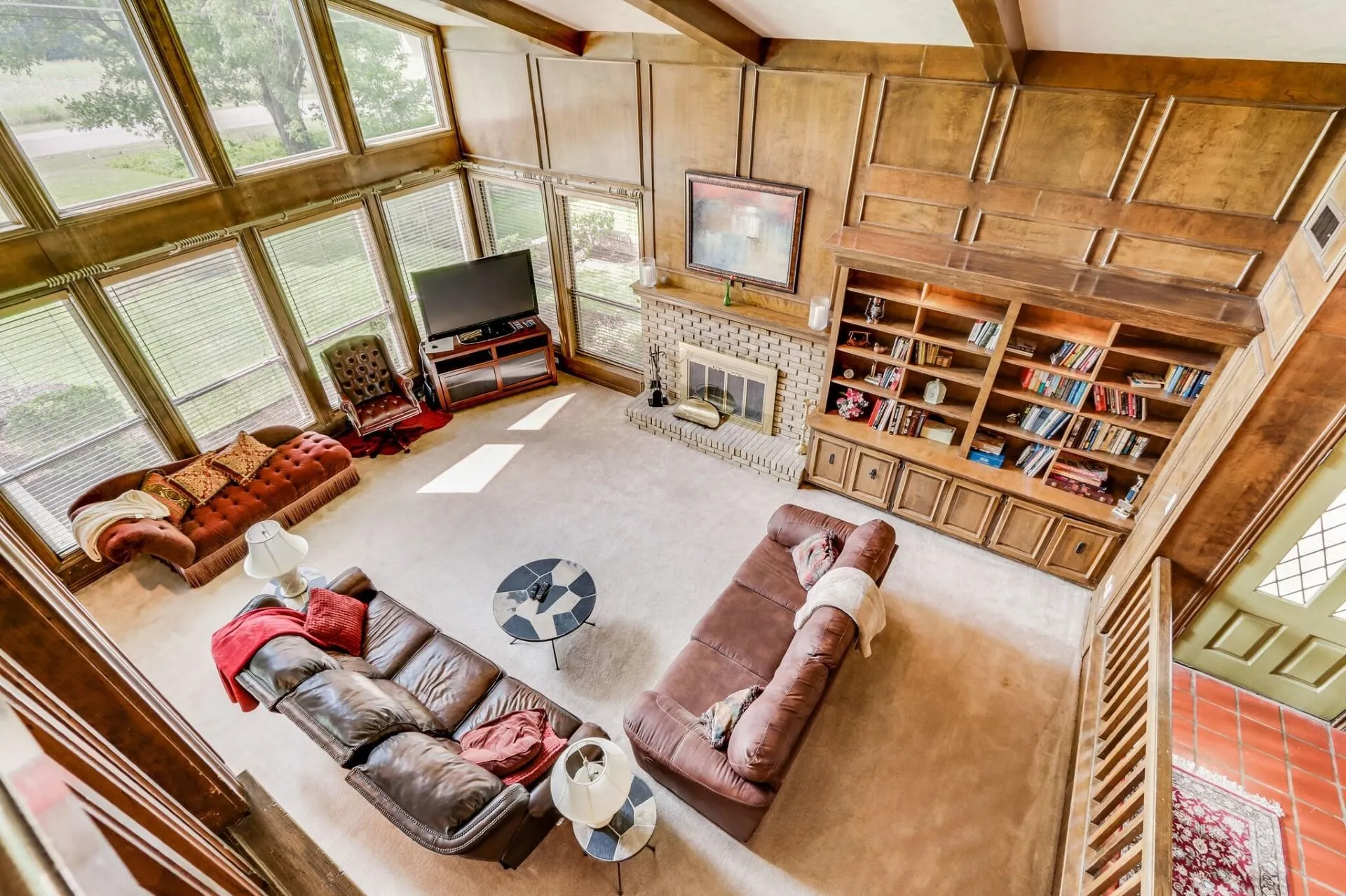 Vaulted living space featuring an exposed beam ceiling, extensive wood paneling, a full-height stone fireplace, built-in shelving, and multiple large windows