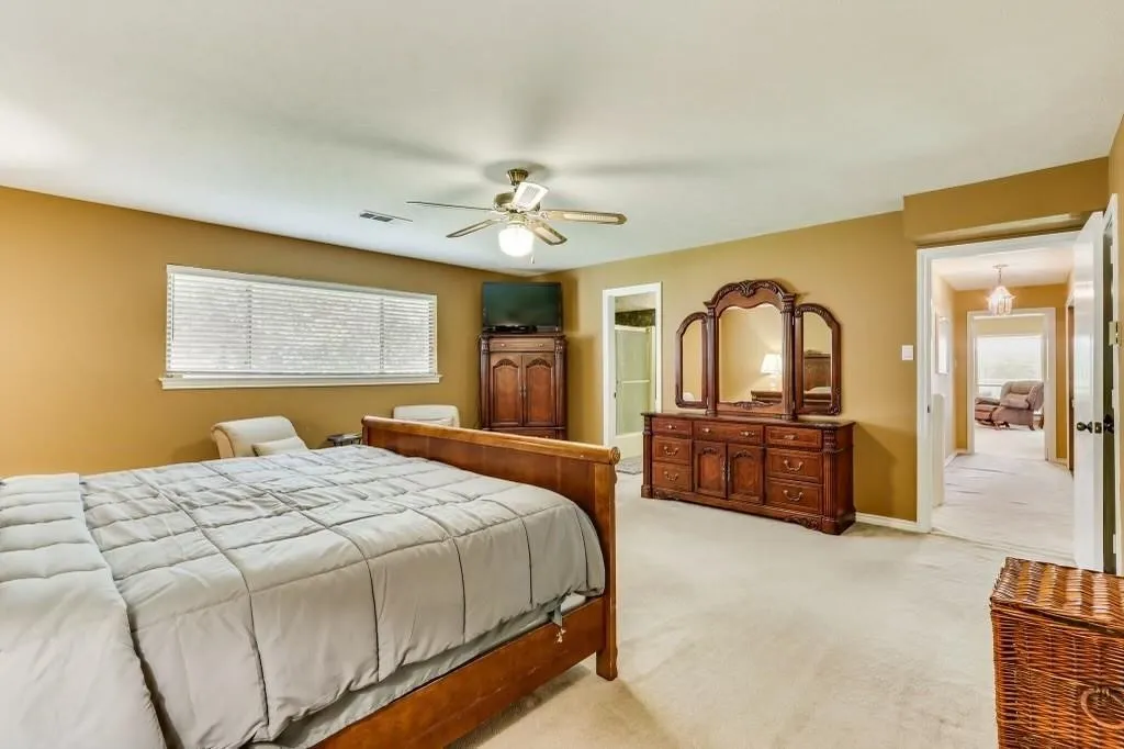 Carpeted bedroom with a single window featuring horizontal blinds, a ceiling fan with integrated lighting, and an open doorway revealing a hall beyond