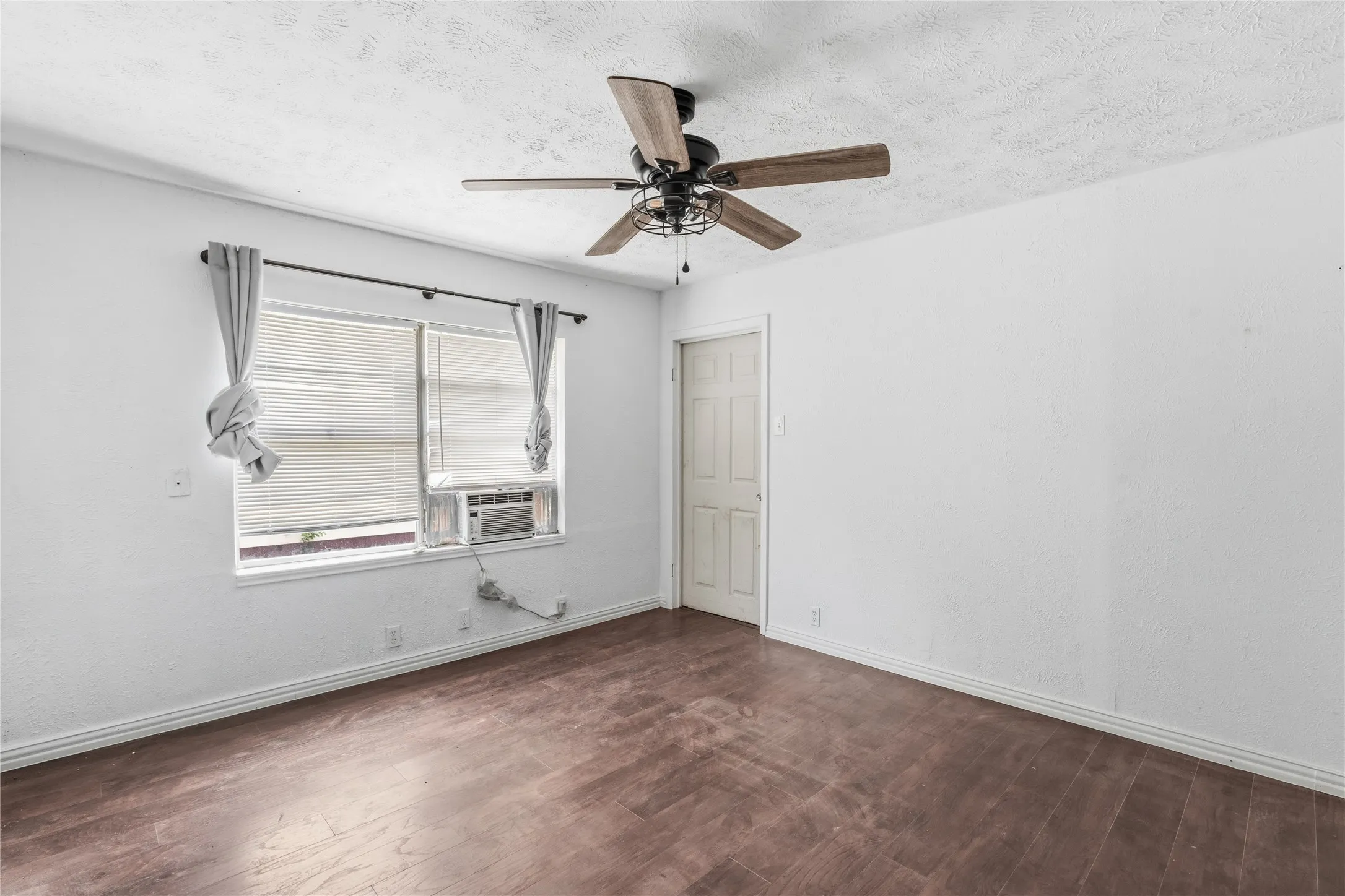 Room featuring wood-finish flooring, a ceiling fan with light fixture, white walls, and a window with blinds and a window-mounted air conditioning unit