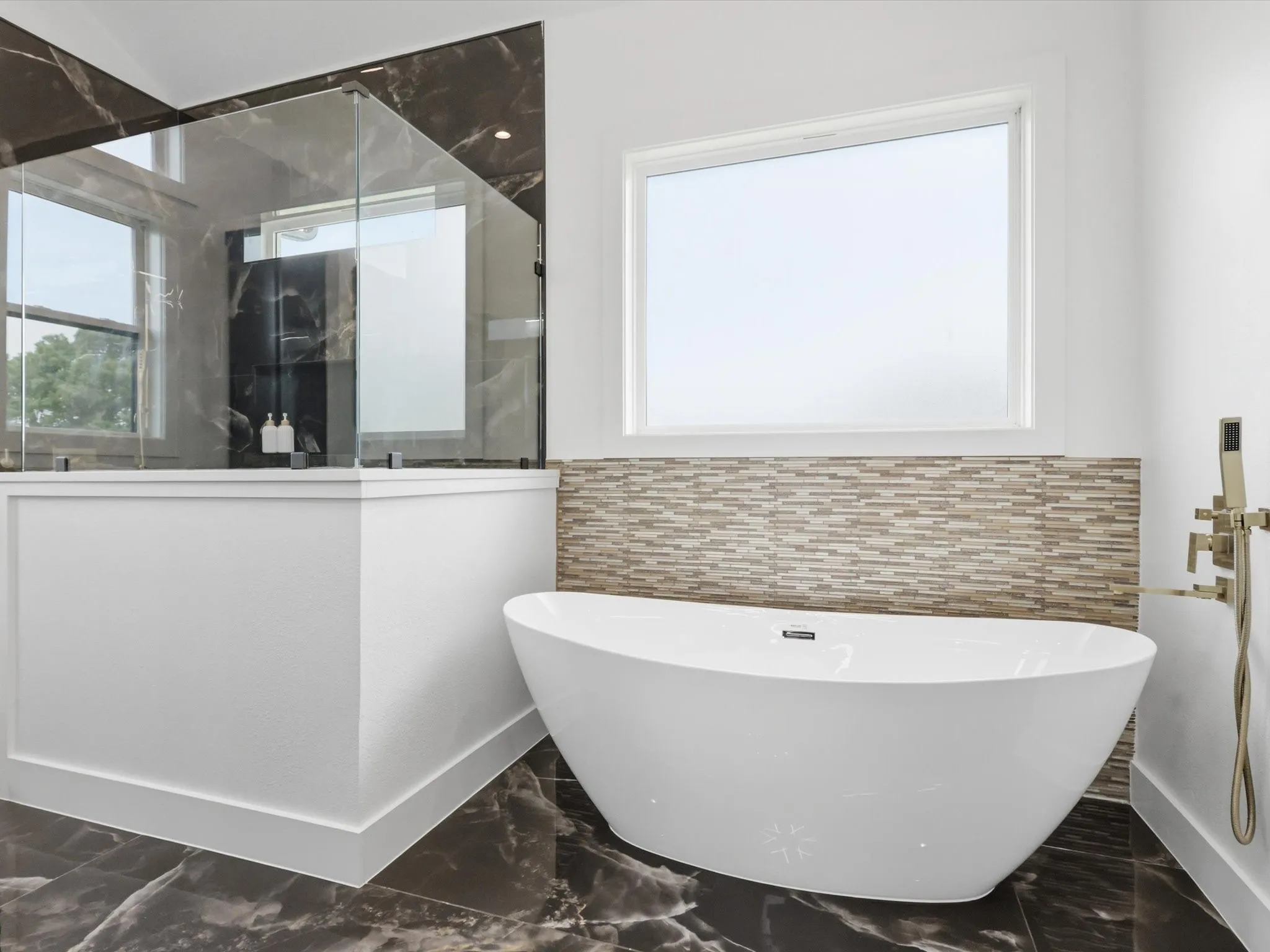 Contemporary bathroom featuring a freestanding soaking tub, a separate glass-enclosed shower with dark marble-look tile, dark marble-look floor tile, and a light-toned mosaic tile accent wall