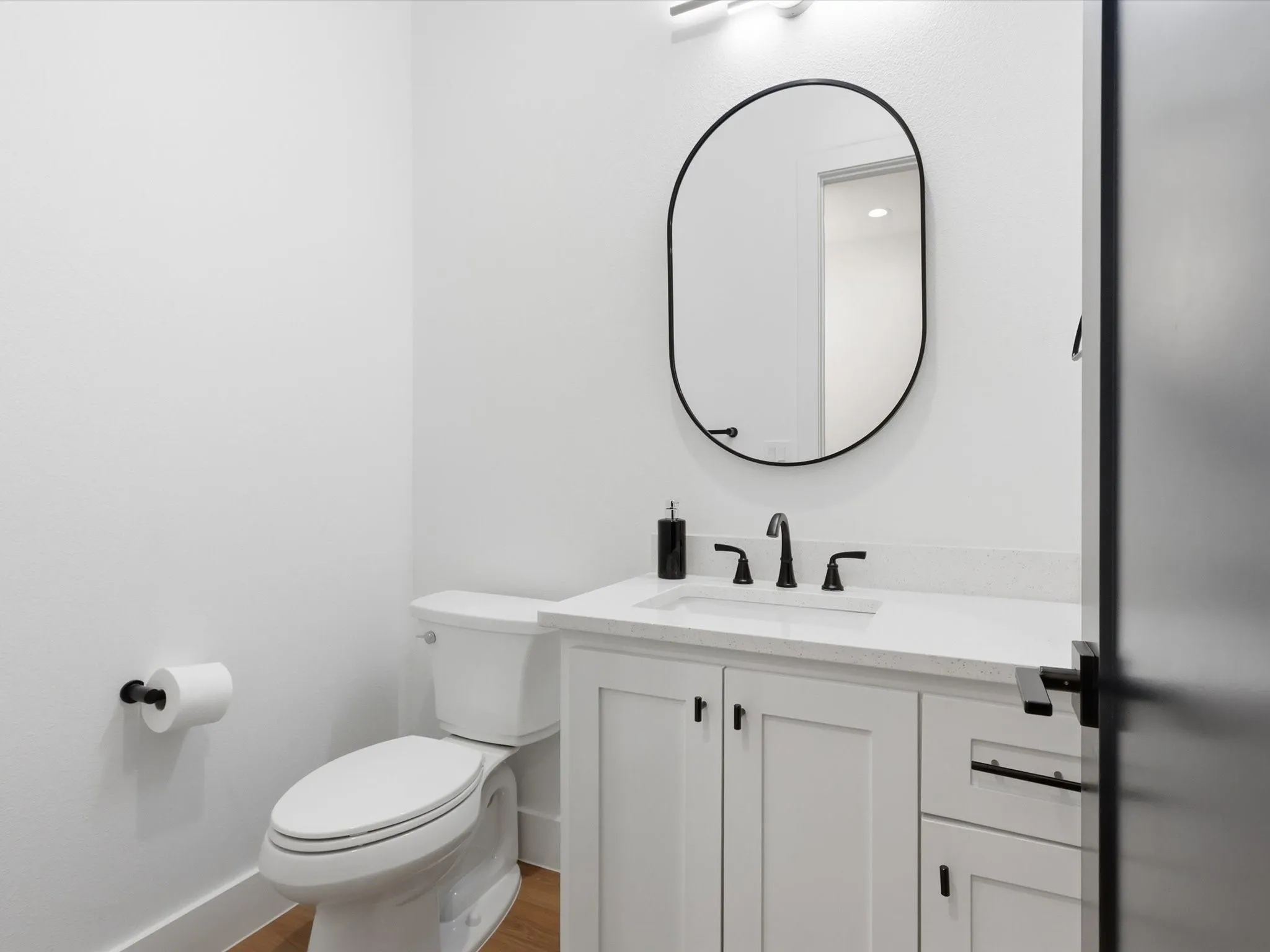 Powder room located on first level, featuring white shaker-style cabinetry, a white countertop with an integrated sink, matte black faucet, and an oval mirror