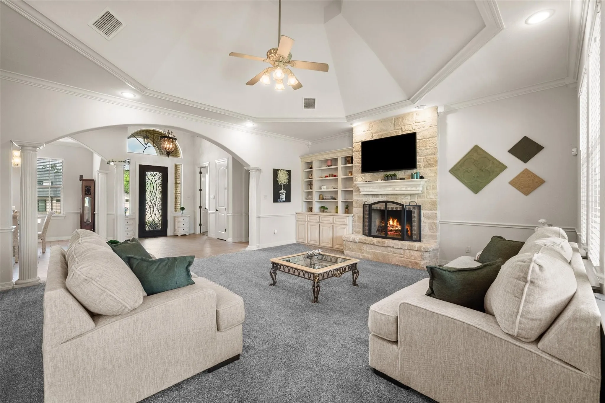 Spacious living area featuring a vaulted ceiling with a fan, a stone-clad fireplace with a mantle, built-in shelving, recessed lighting, and large windows with blinds