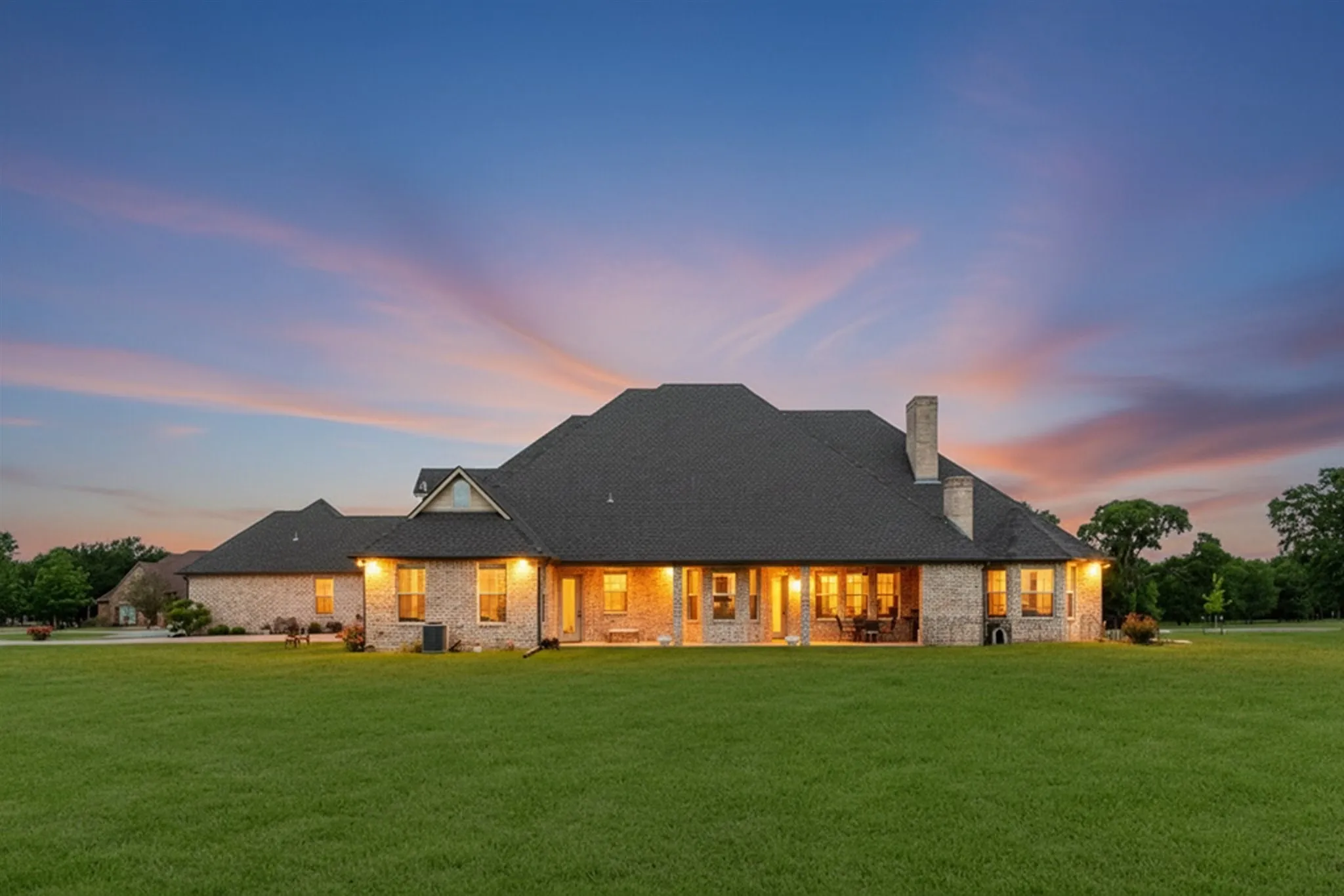 Expansive green lawn with a sprawling brick exterior residence featuring multiple windows, a covered patio, and a shingled roof