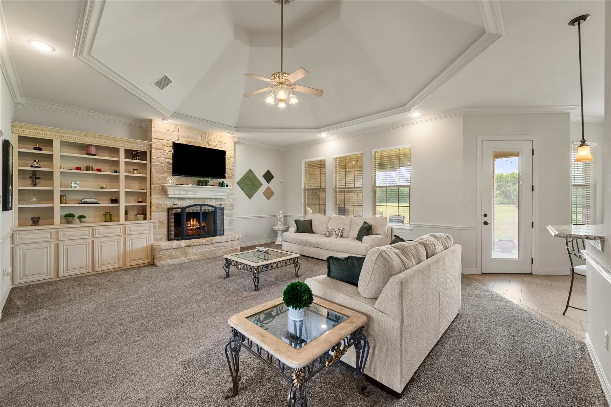 Living area featuring a tray ceiling with a ceiling fan, a stone fireplace with a hearth, built-in shelving with lower cabinetry, and multiple windows
