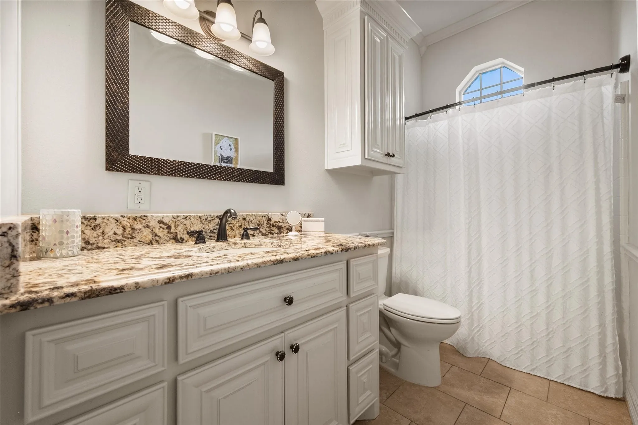 Ensuite bathroom featuring a white vanity with granite countertop, a framed mirror, a built-in wall cabinet, and a window with an arched transom