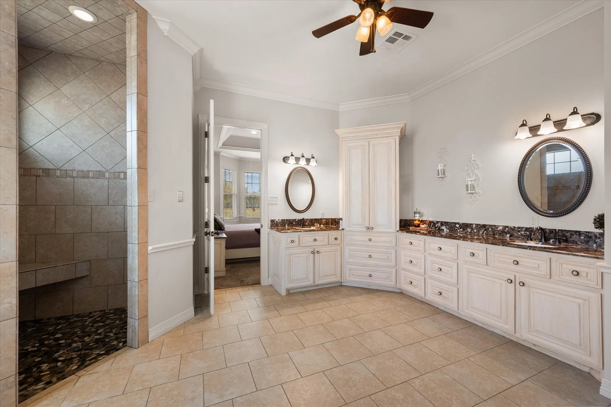 Spacious bath featuring a tiled walk-in shower with built-in bench, dual vanities with dark countertops and integrated sinks, marble countertops, extensive cabinetry storage, and tile flooring