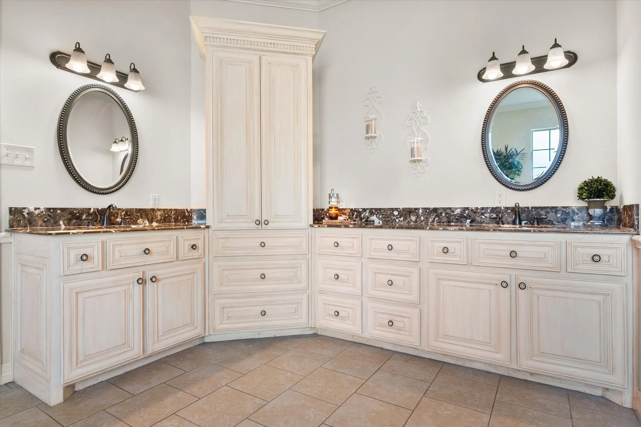 Dual vanities with undermount sinks, flanked by extensive white cabinetry, dark marble countertops, and complementing oval mirrors with wall-mounted light fixtures
