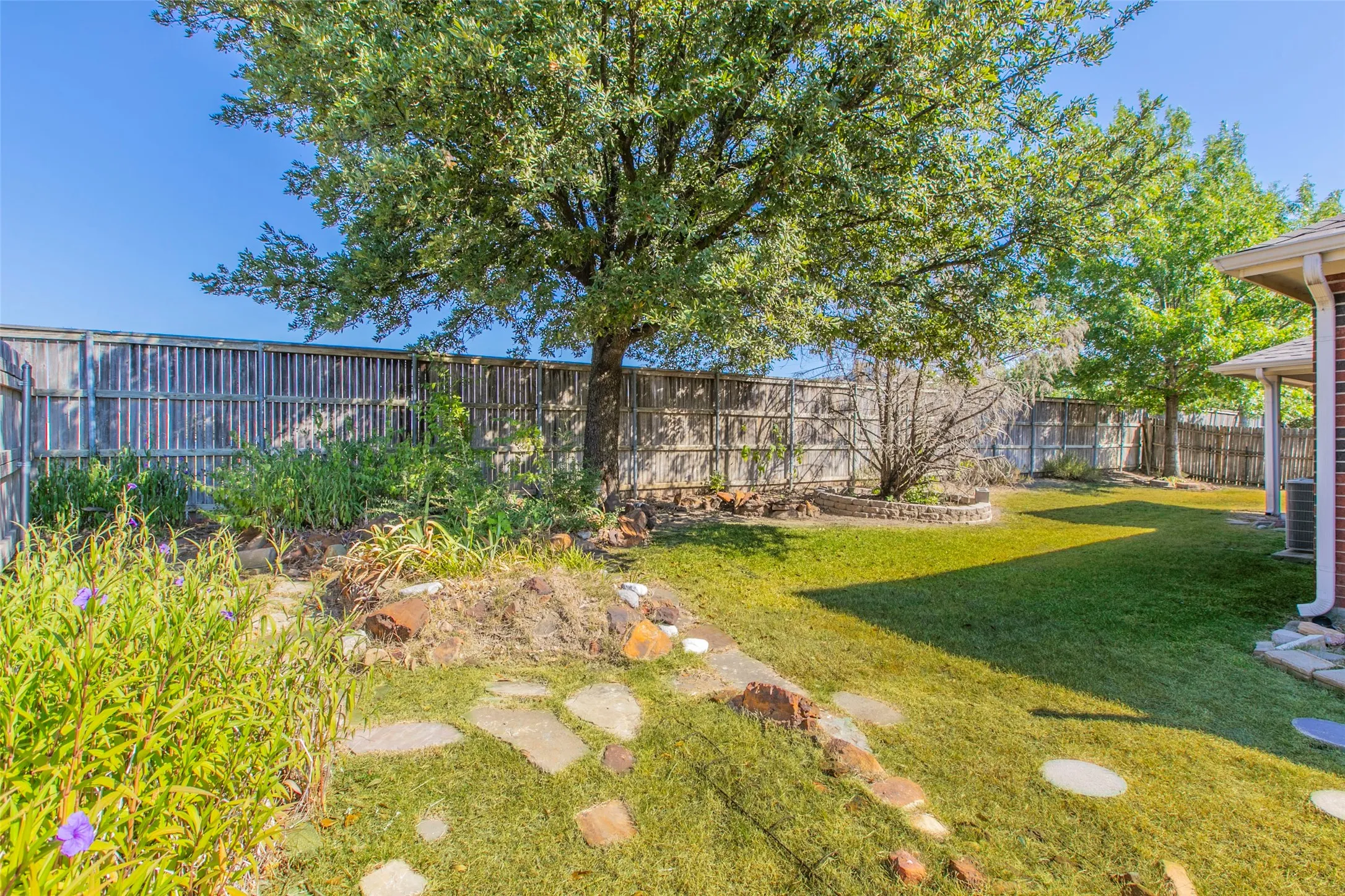 Expansive backyard featuring a manicured lawn, established trees, a raised garden bed with stone retaining walls, and stepping stones