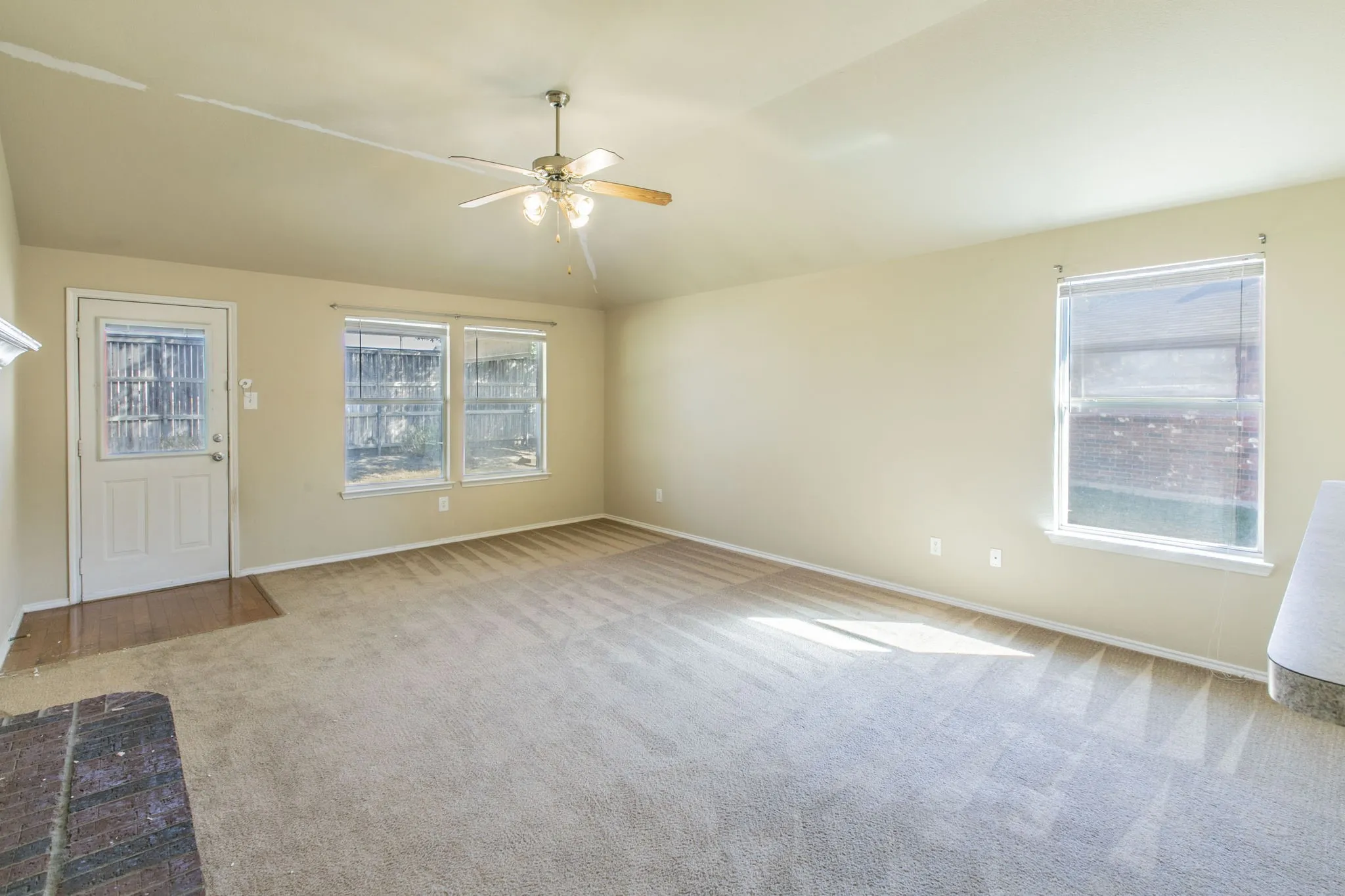 Spacious carpeted room featuring a ceiling fan, multiple windows, a rear exterior door, and a fireplace hearth with stone-look tile