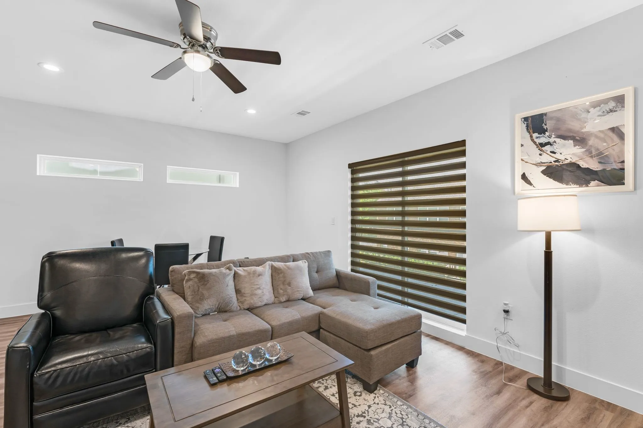 Living area featuring hardwood style flooring, a large window with horizontal blinds, and a ceiling fan with integrated lighting