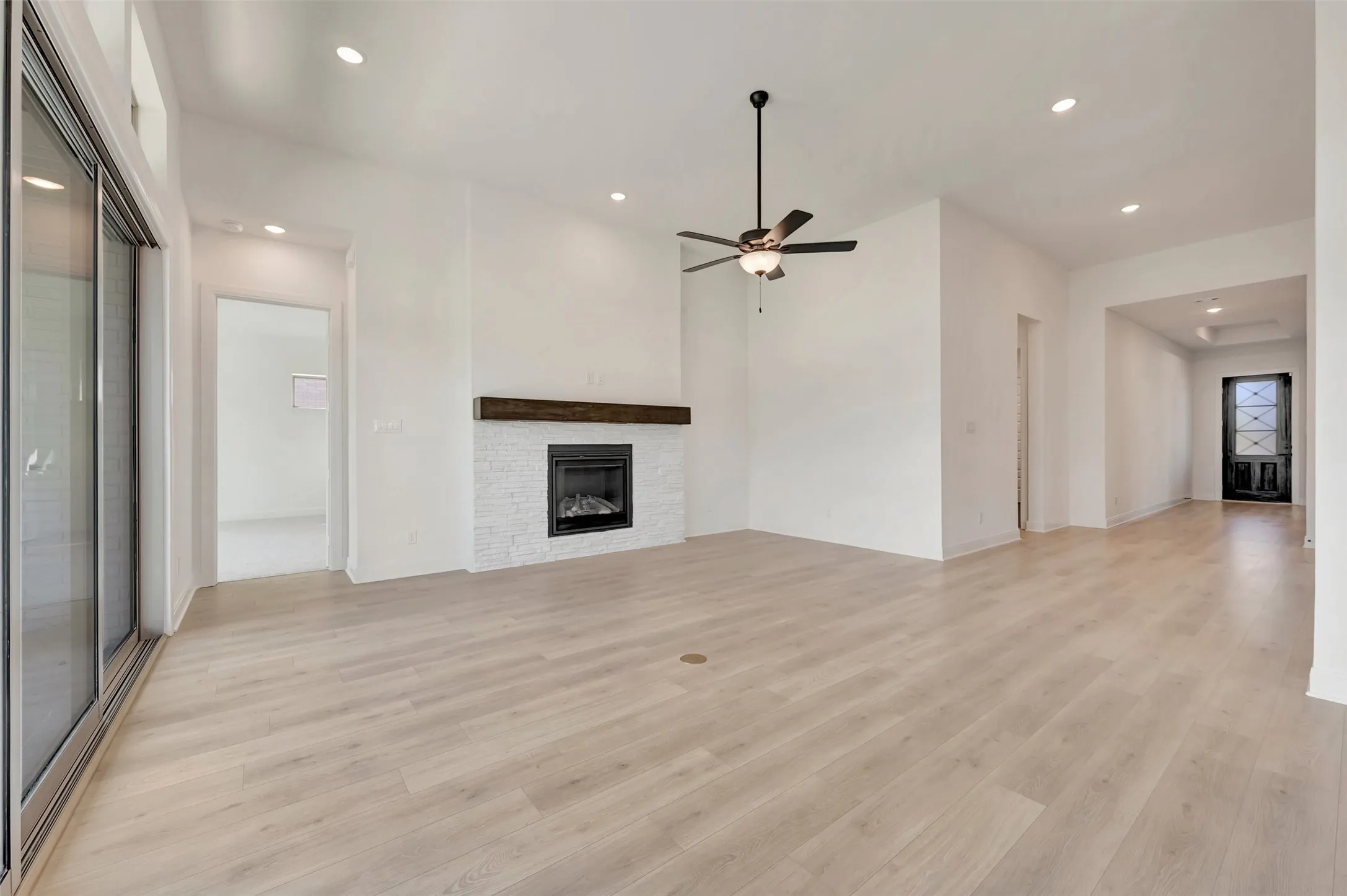 Spacious living area featuring light wood-finish flooring, a contemporary fireplace with white brick-style surround and dark wood mantel, and a dark-toned ceiling fan
