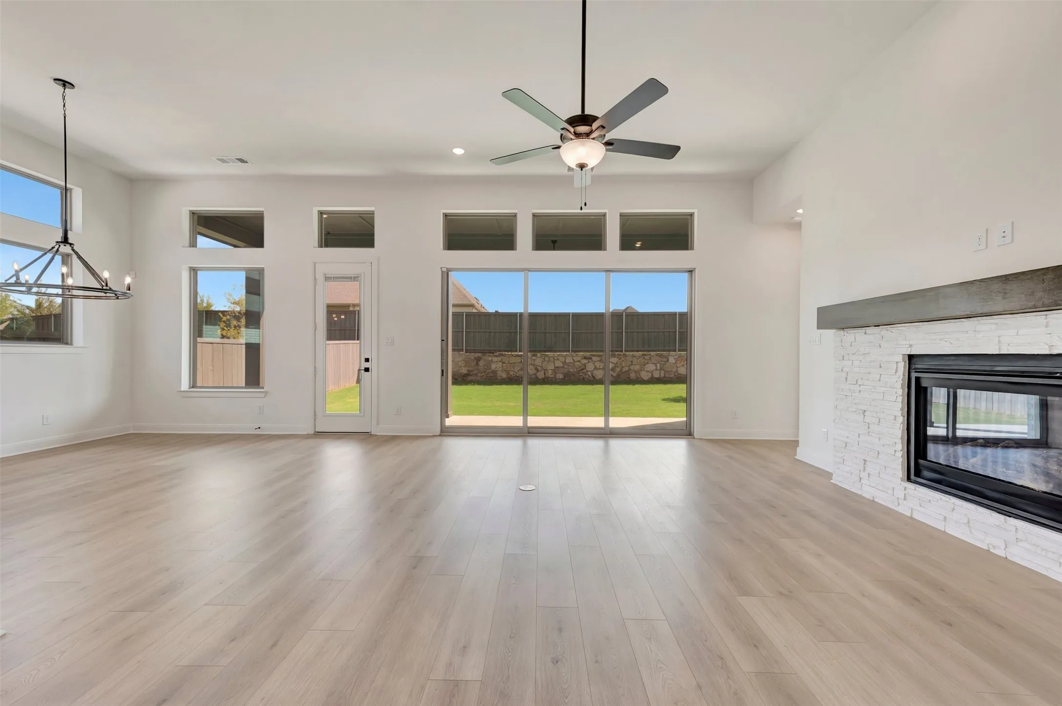 Spacious living area featuring light wood-finish flooring, a contemporary fireplace with white brick surround and a concrete mantel, a ceiling fan, and extensive natural light from multiple windows and a sliding glass door leading to the backyard