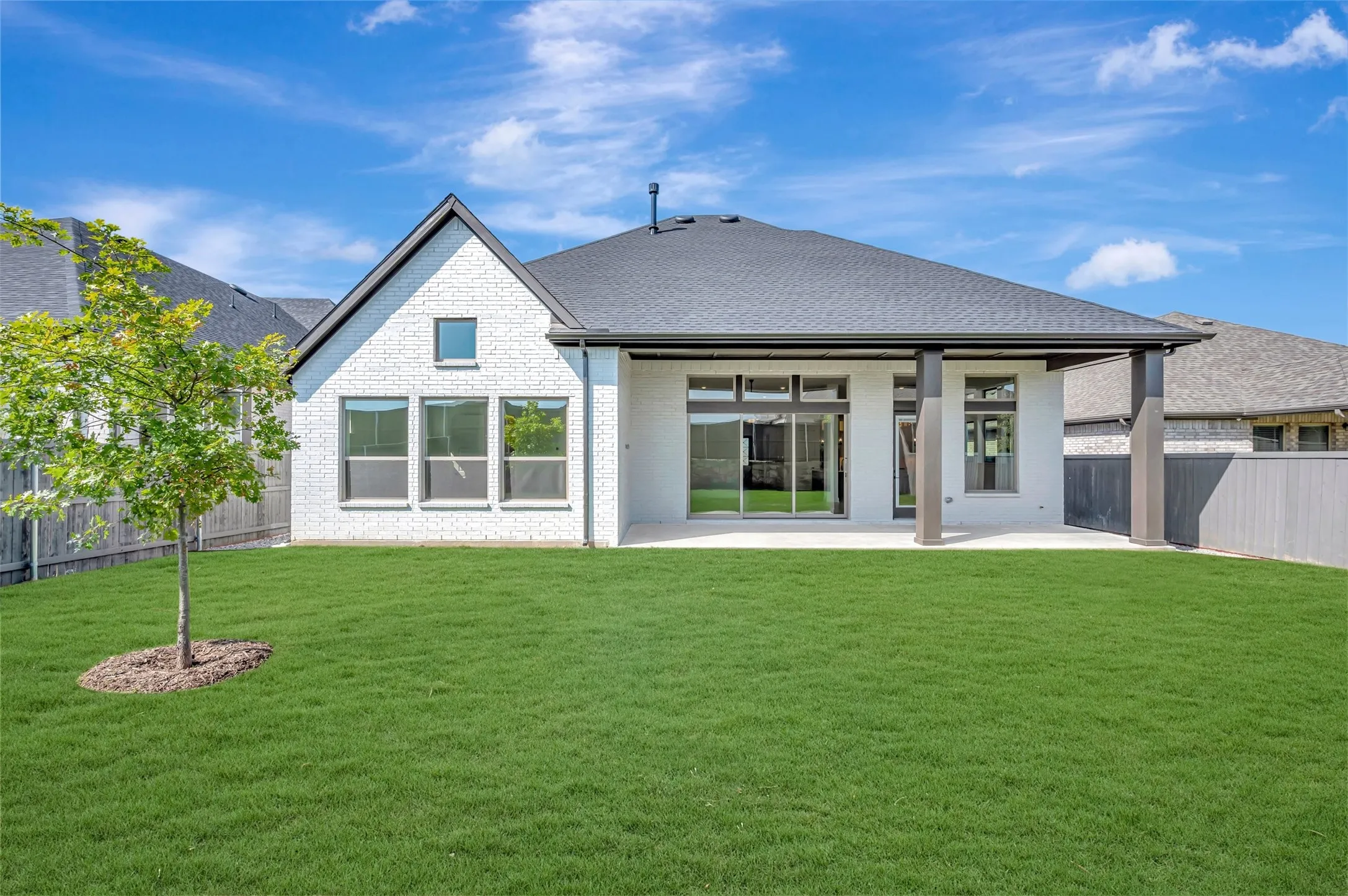 Expansive rear exterior featuring a white brick facade, dark roofing, a covered patio with dark support columns, multiple windows, and a generous lawn space