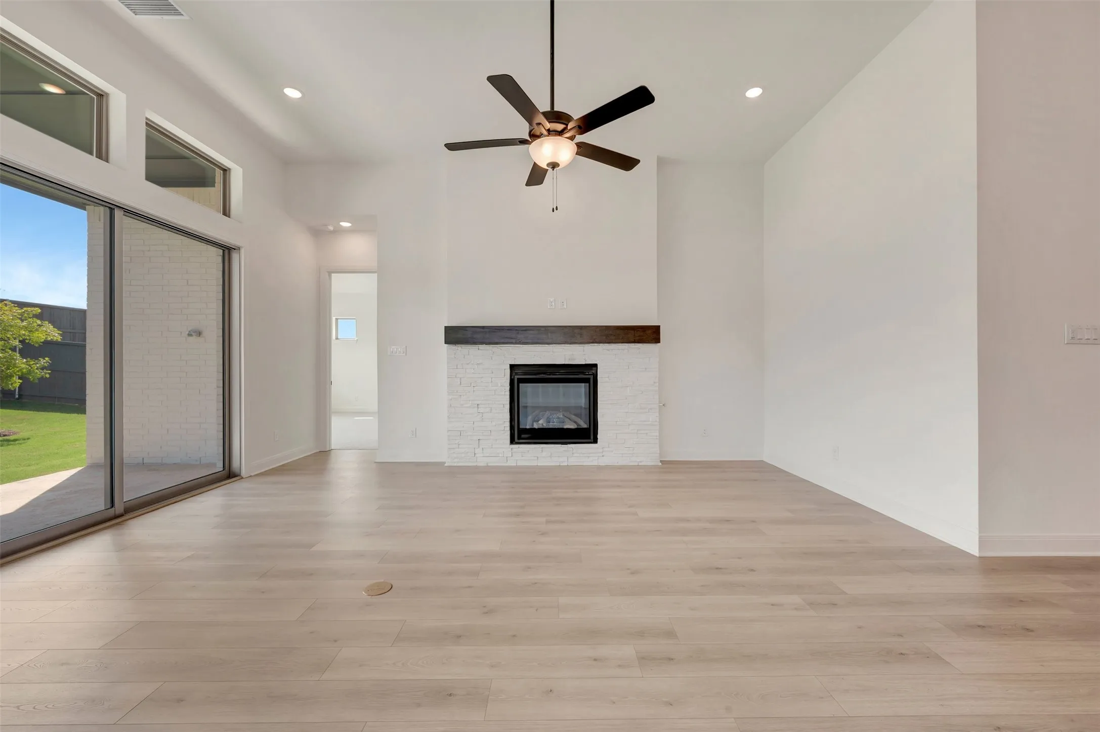 Spacious interior featuring wood-finish flooring, a contemporary fireplace with a white brick surround and dark wood mantel, and a ceiling fan with integrated lighting