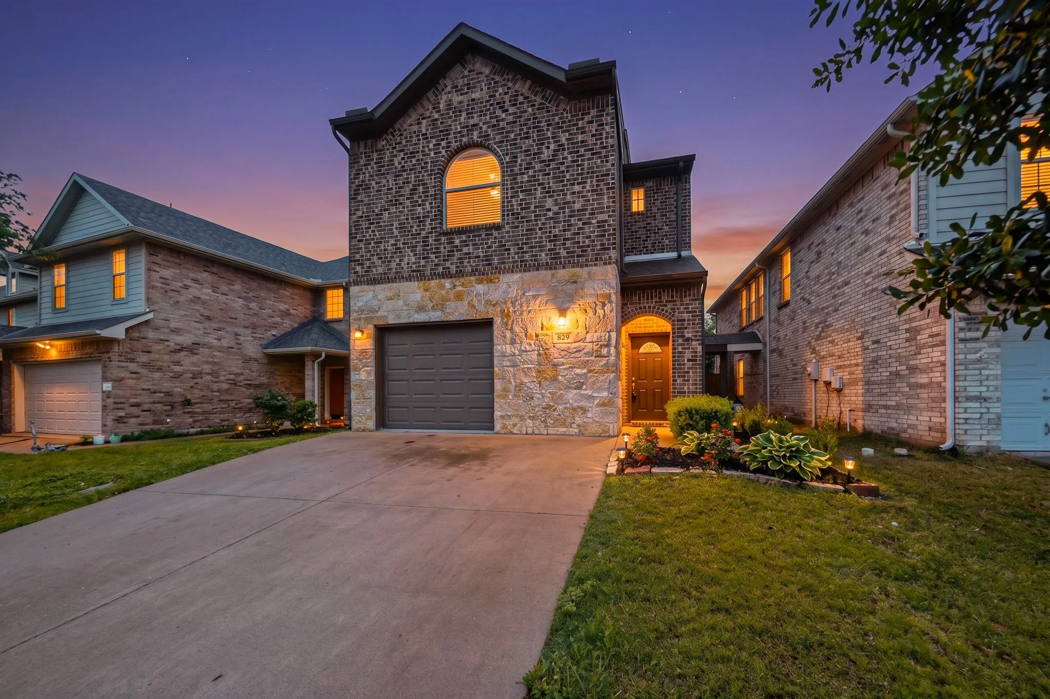Two-story residence featuring a brick and stone facade, arched entryway, an integrated garage, a concrete driveway, and established landscaping