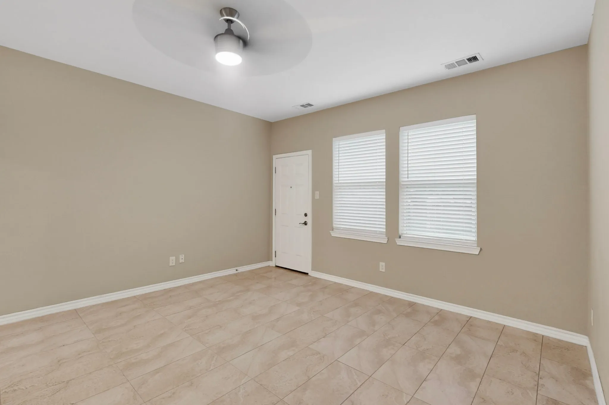 Spacious room featuring neutral wall paint, tile flooring, two windows with blinds, a white entry door, and a ceiling fan with integrated lighting