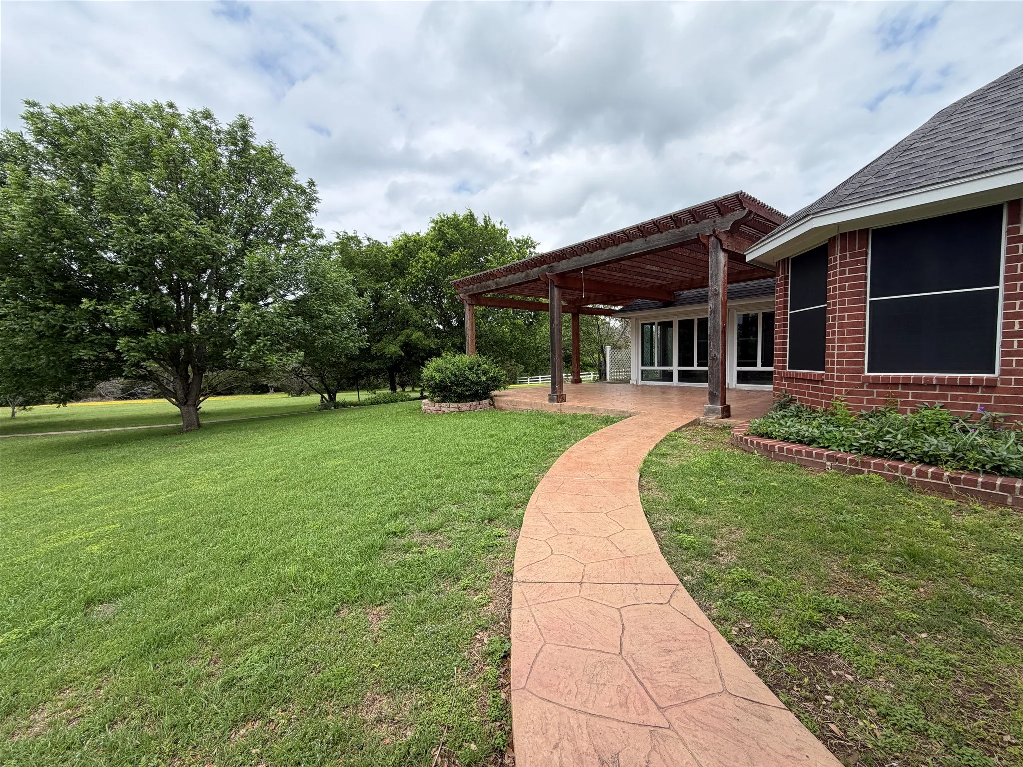 Expansive rear yard featuring a stamped concrete pathway, mature trees, and a covered patio with a wooden pergola