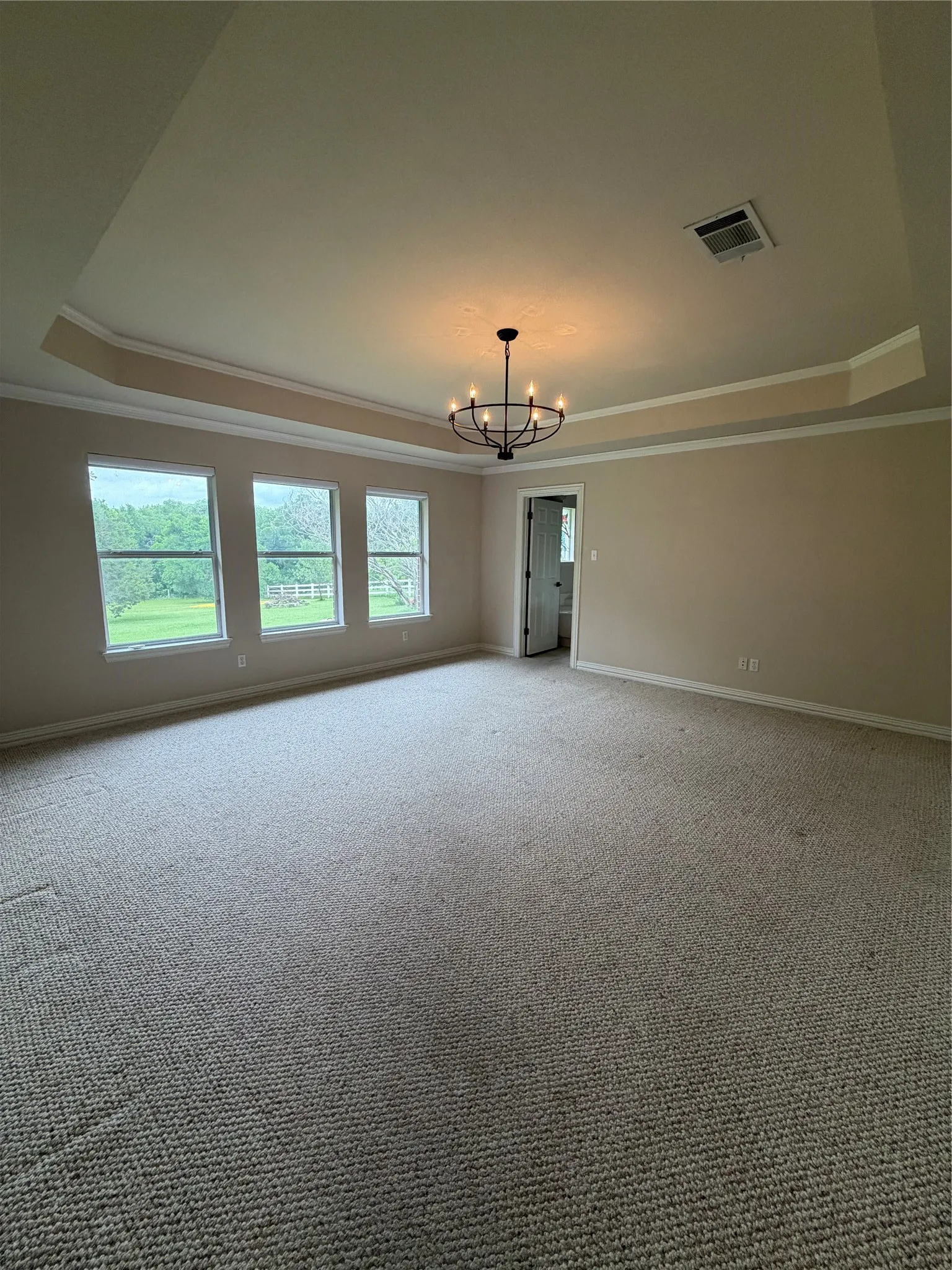 Spacious room featuring neutral carpet flooring, three large windows, a tray ceiling with crown molding, and a black metal chandelier