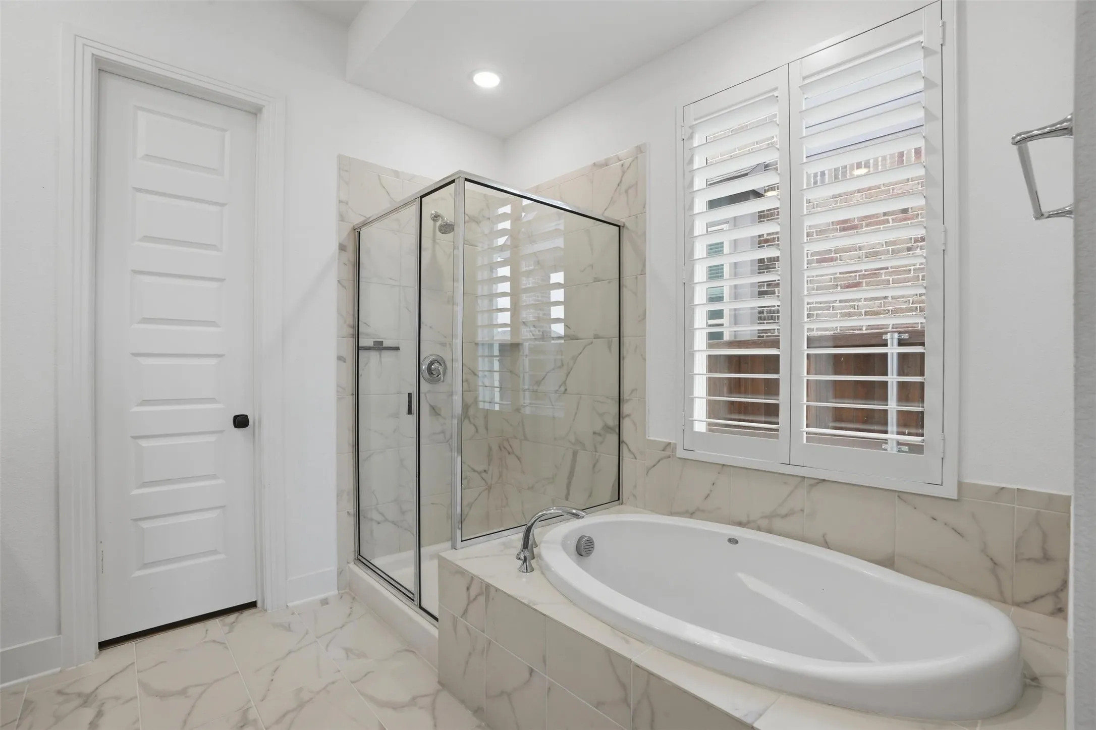 White paneled door opening into a bathroom featuring a frameless glass shower enclosure, a built-in soaking tub, plantation shutters, and large-format porcelain tile flooring and wainscoting