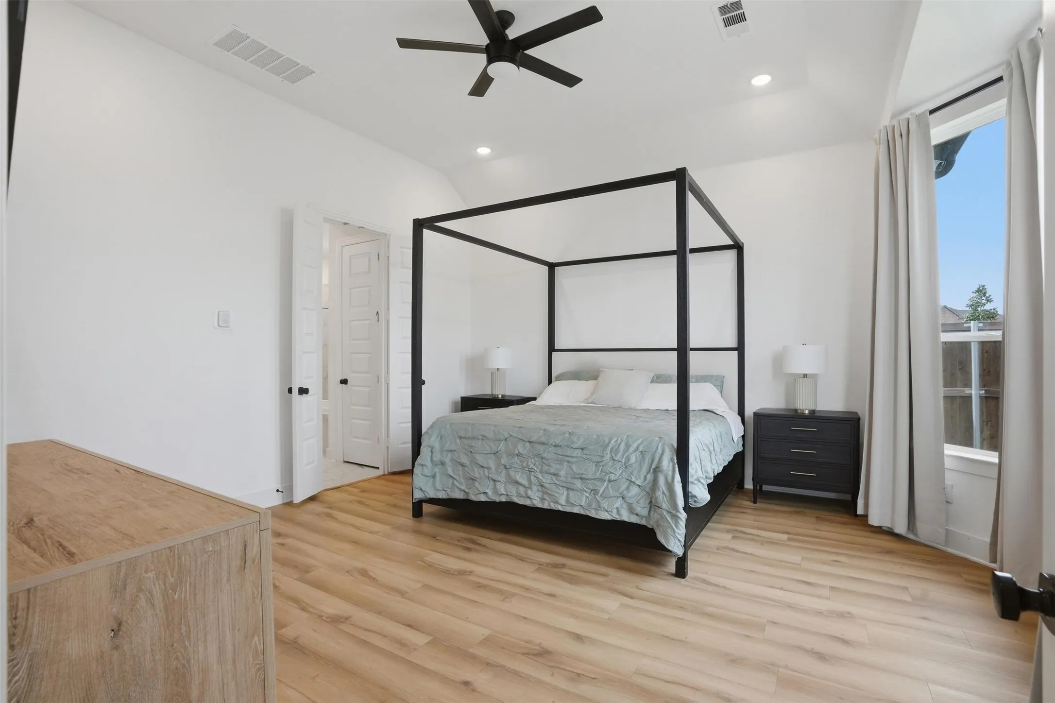 Spacious room featuring light wood-finish flooring, a vaulted ceiling, a ceiling fan with integrated lighting, recessed lighting, and a large window with drapes
