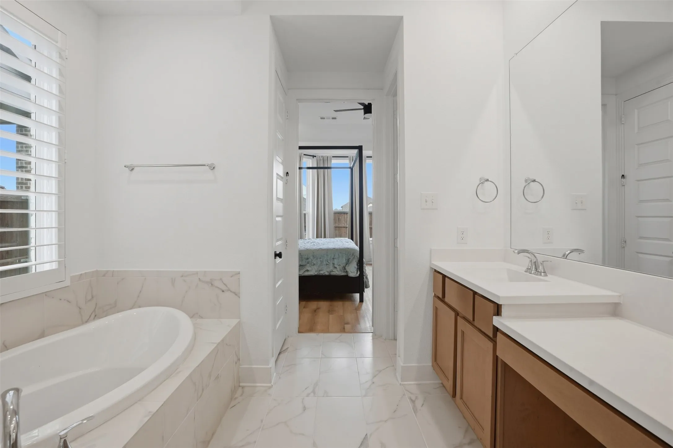 Bathroom featuring a freestanding tub with marble-look surround, white plantation shutters, vanity with light wood cabinetry and white countertops, and expansive wall mirror