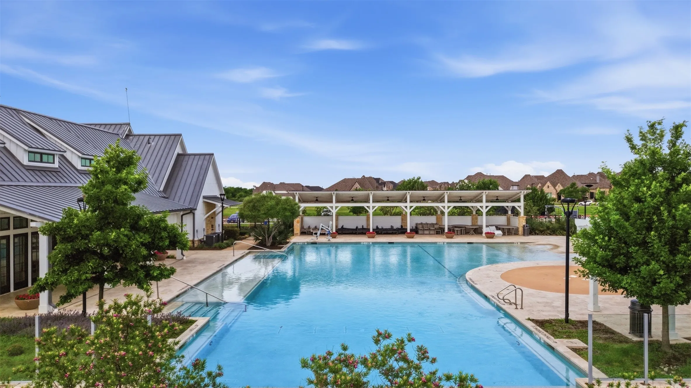 Community pool area featuring a large swimming pool, expansive paved sun deck, covered cabana with multiple seating areas, and a classic-style clubhouse with a metal roof