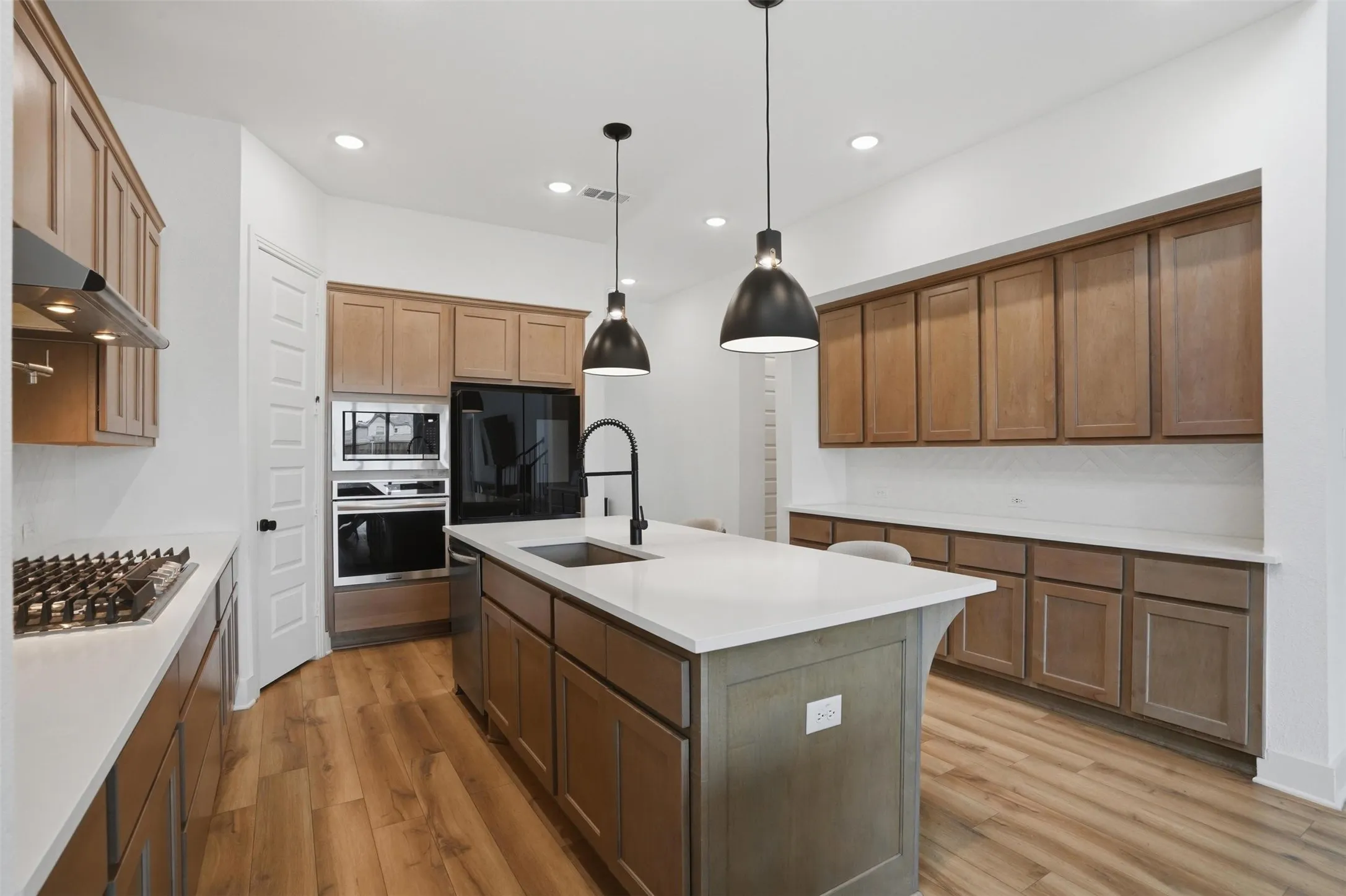 Modern kitchen featuring a central island with white countertop, black pendant lighting, wood-finish flooring, built-in stainless steel appliances, and extensive cabinetry