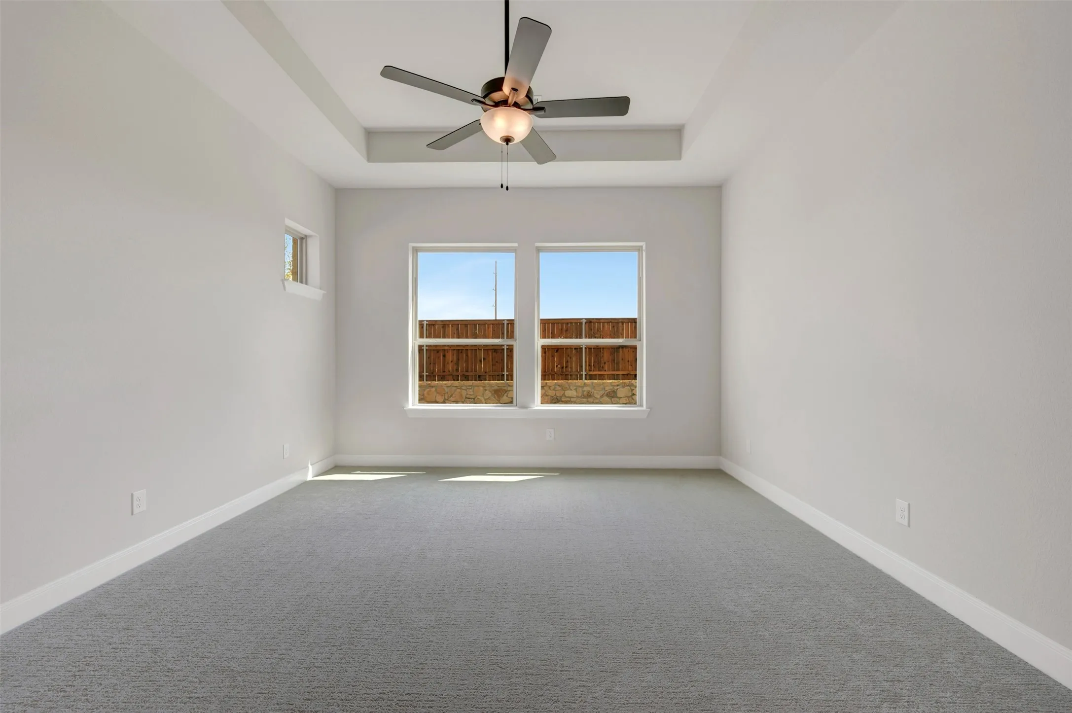 Tray ceiling with integrated ceiling fan, two large windows, and a smaller casement window providing ample natural light, and grey carpeting