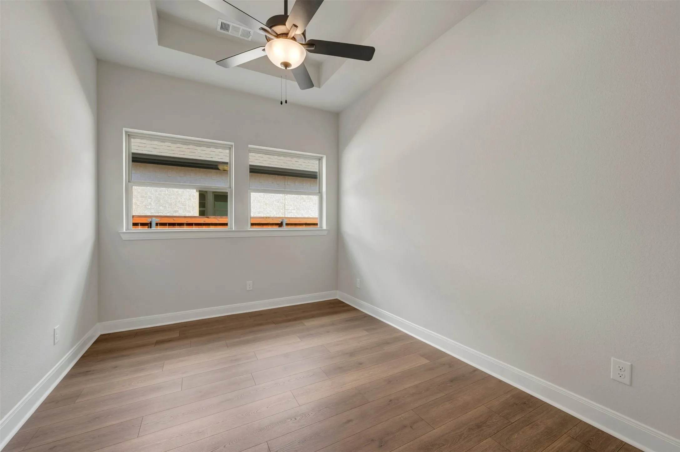 Wood-finish flooring, white baseboards, two windows with white trim, a tray ceiling, and a ceiling fan with integrated lighting