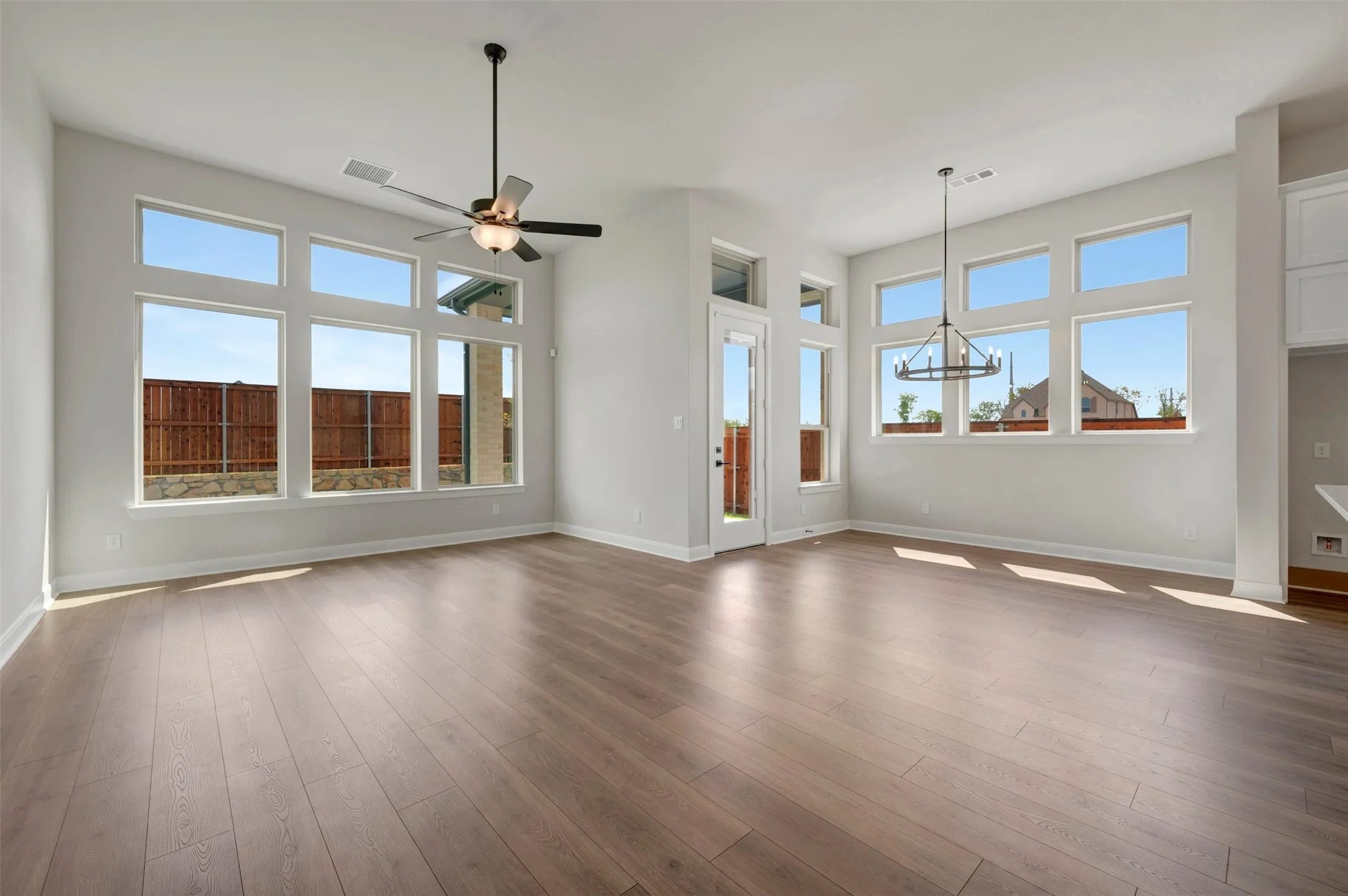Expansive living space featuring wood-finish flooring, abundant natural light from multiple windows, a ceiling fan with integrated lighting, and a contemporary chandelier
