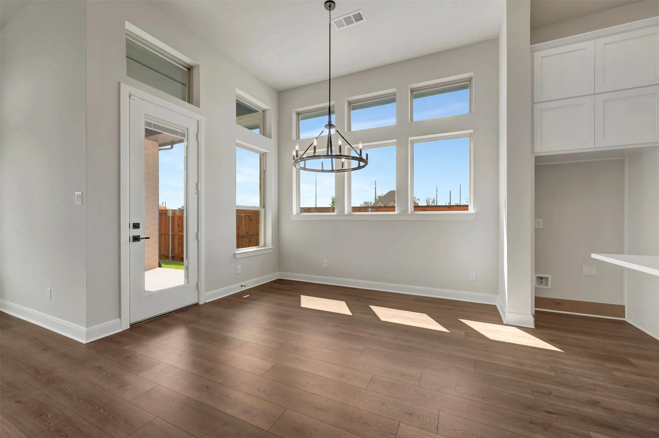 Expansive wood-finish flooring, a contemporary chandelier, abundant natural light from multiple windows, a glass-paneled door, and built-in cabinetry enhance this space