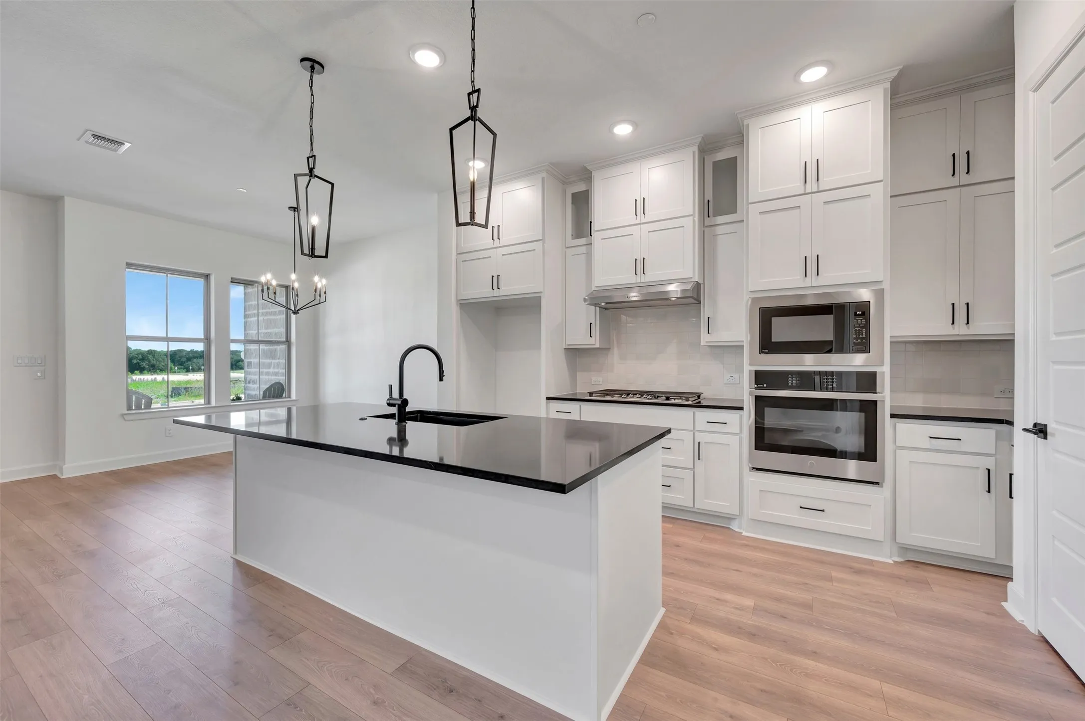 Open-concept kitchen featuring an oversized island with a black sink and faucet, white shaker cabinetry, stainless steel appliances, and light wood-finish flooring
